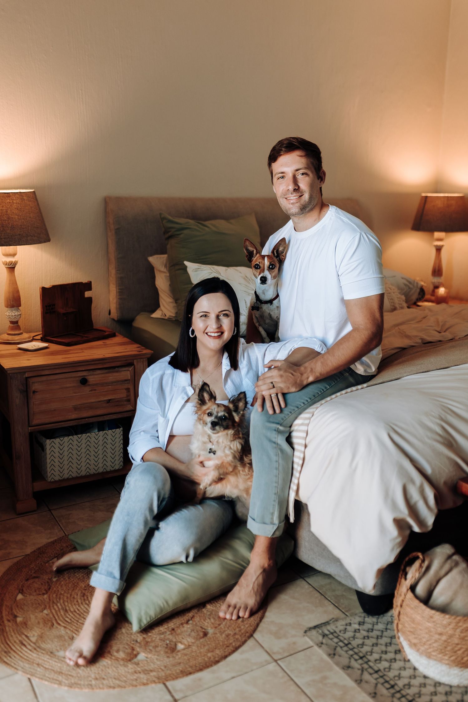 A couple relaxes on the floor with their small dog in a warmly lit bedroom.