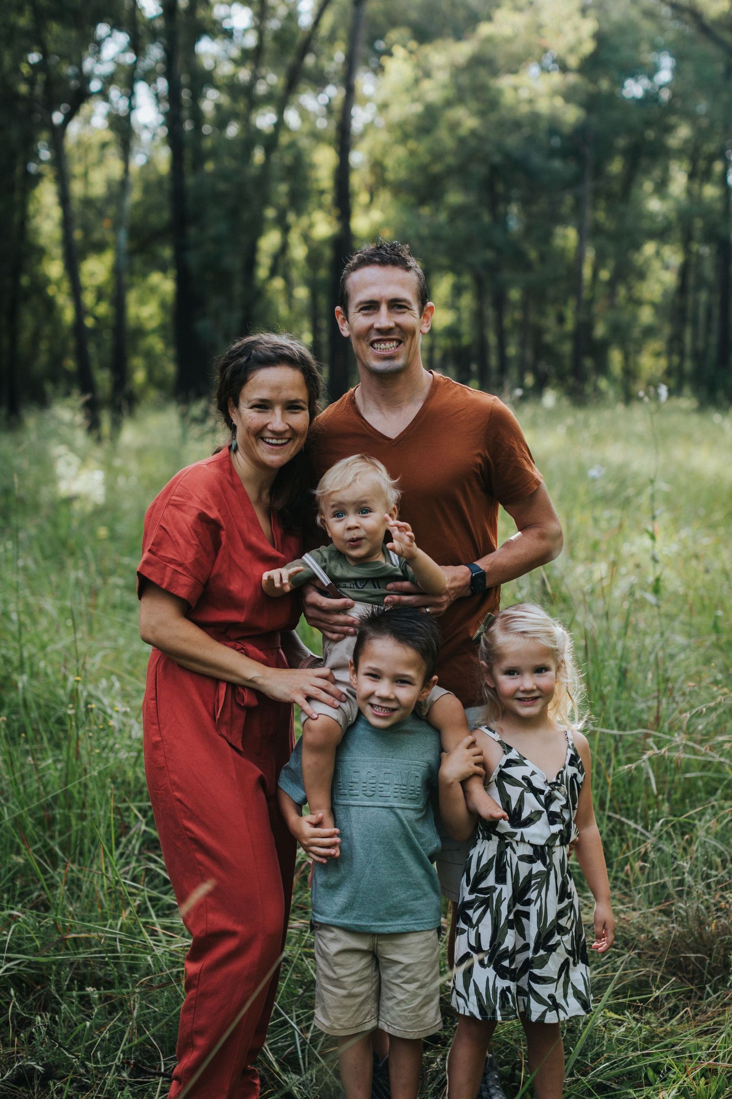 A happy family of five poses together for a portrait in a sunlit forest setting with tall grass and trees.