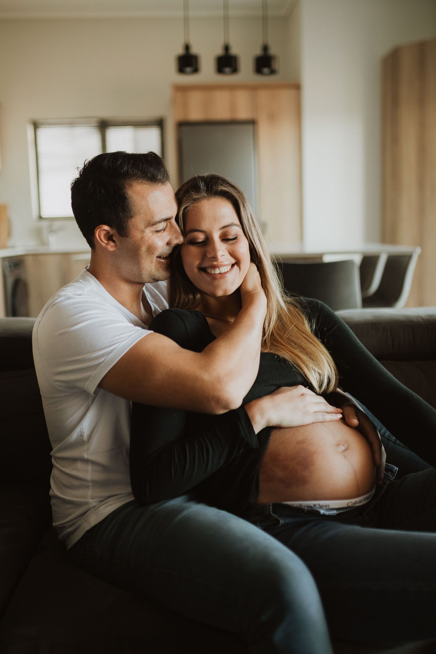Couple shares tender moment together during indoor pregnancy photoshoot on sofa.