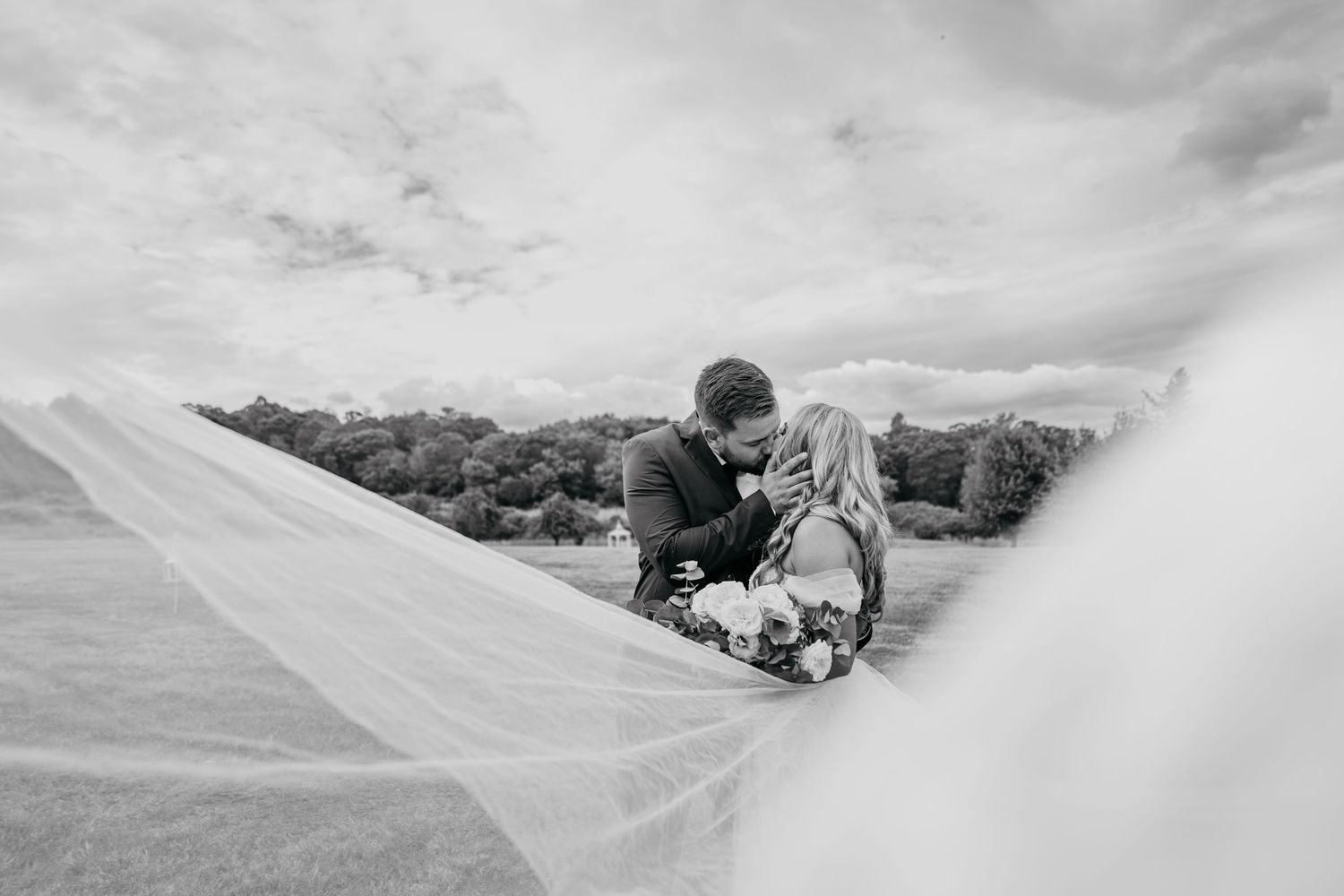 A black and white photo of a person laying down in a flowing white dress against a cloudy sky background.