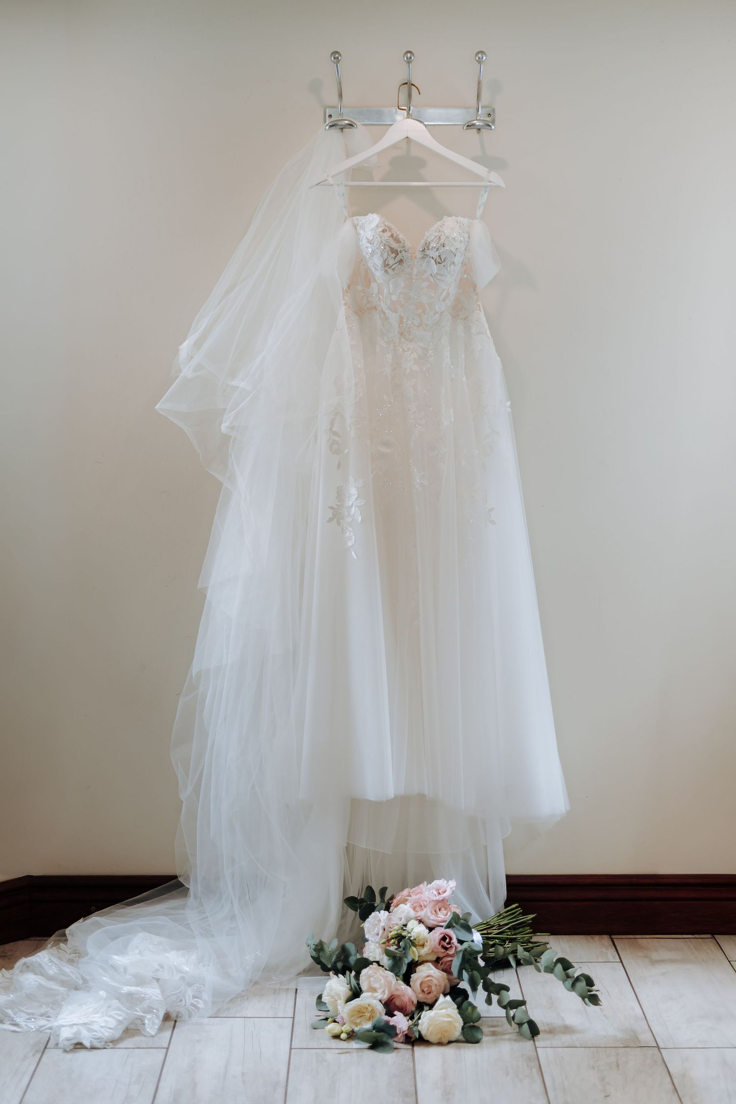 White lace wedding dress with tulle skirt displayed on hangers with soft floral decorations against neutral background.