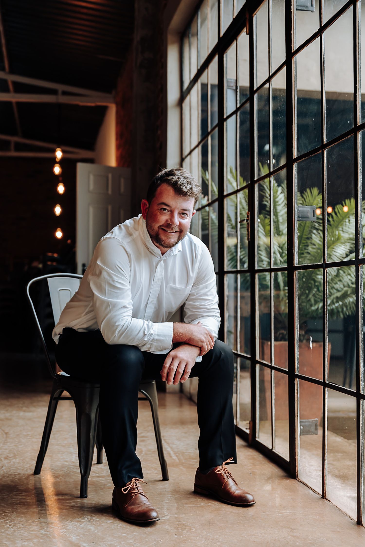 Professional headshots of a man in white shirt and dark pants sitting by large windows in an industrial style space.