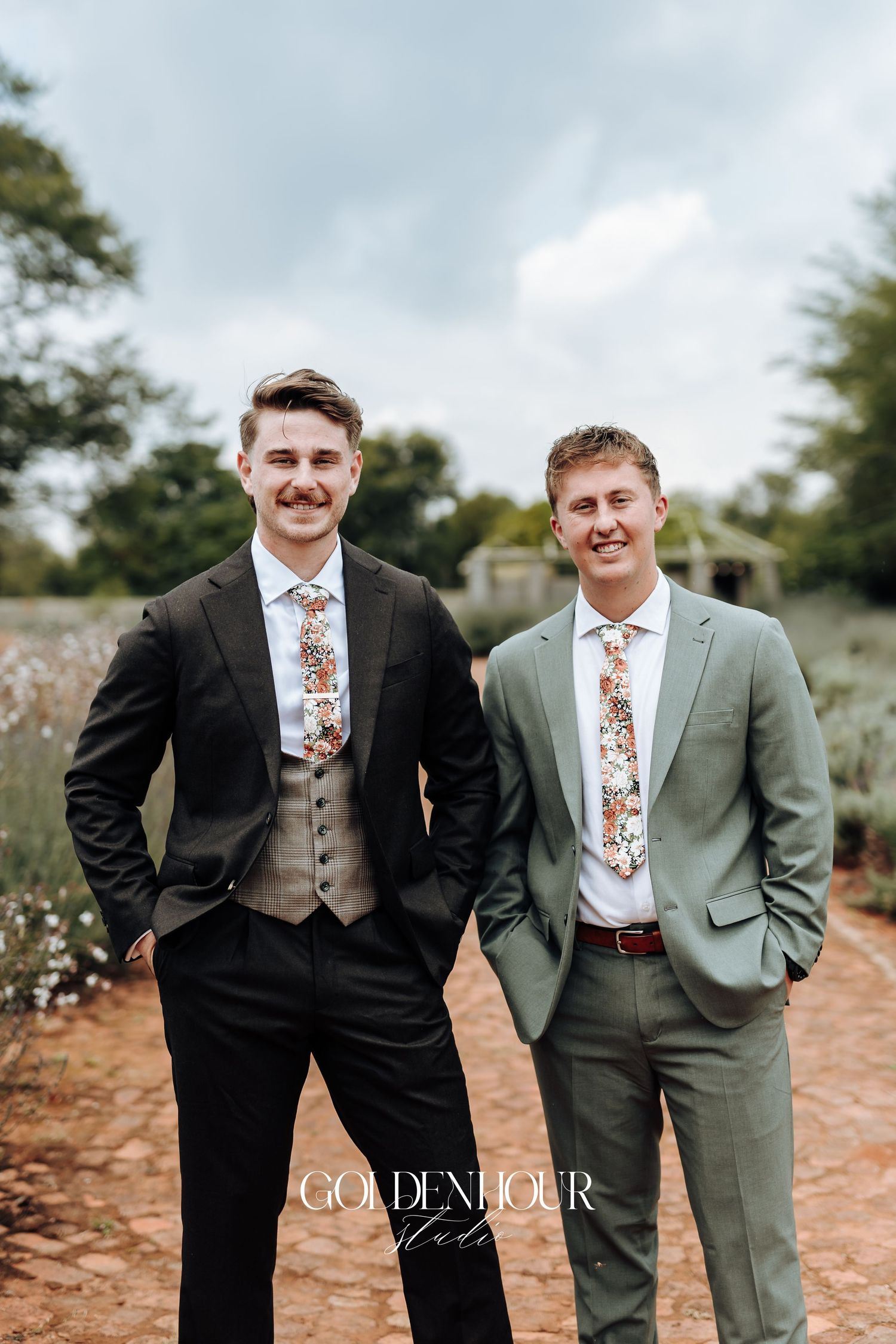 Two groomsmen in contrasting black and sage green suits pose together outdoors against a natural backdrop.