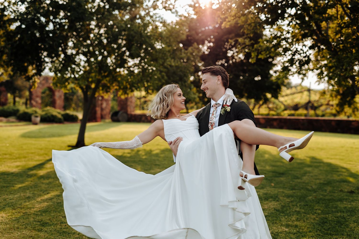A couple in wedding attire dances together in a sunlit field during golden hour.