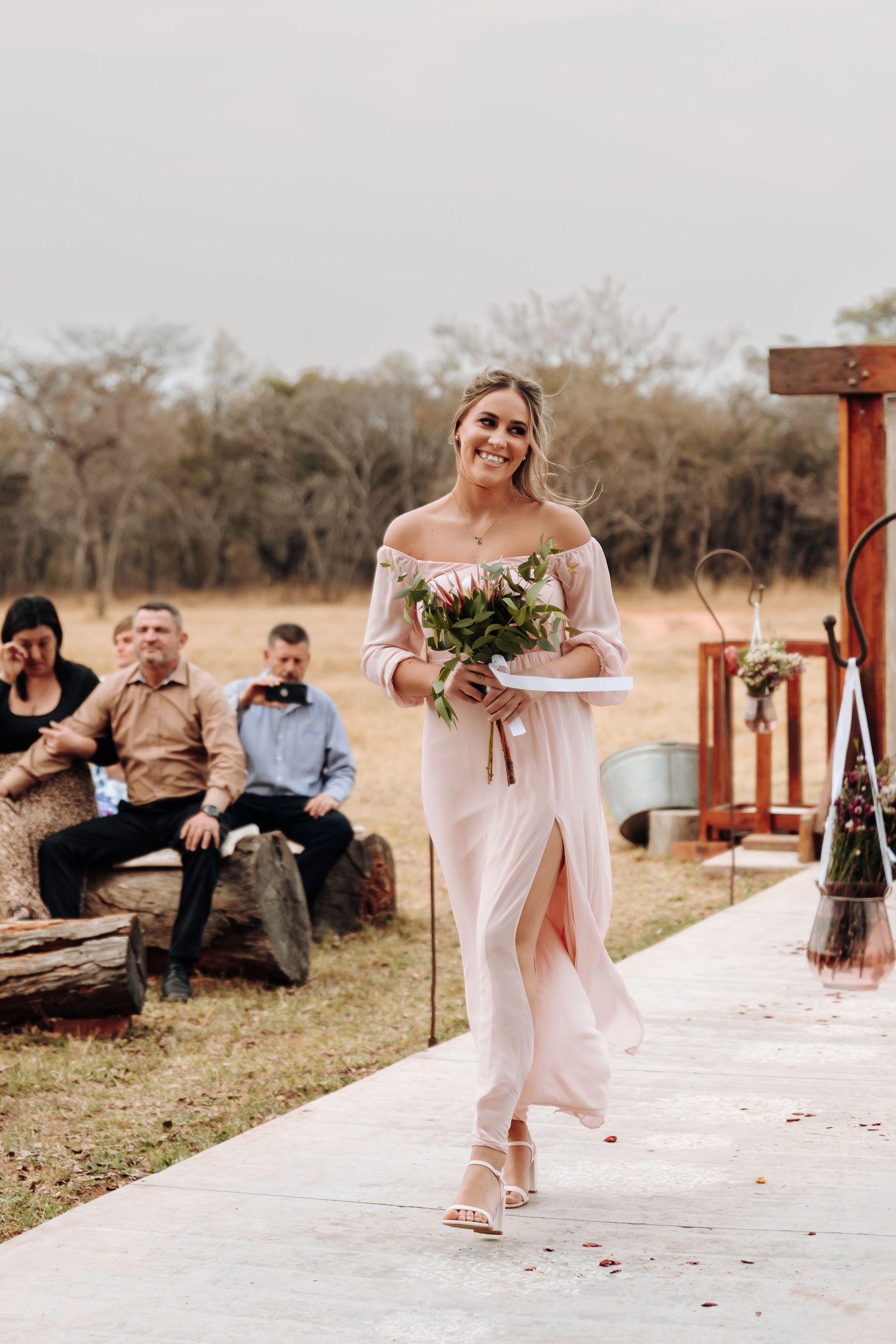 A bride walks down an outdoor aisle in a flowing off-shoulder dress carrying a bouquet at a rustic wedding ceremony.
