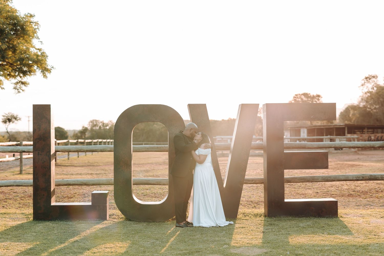 Bride in flowing white dress poses by large LOVE letters during sunset.