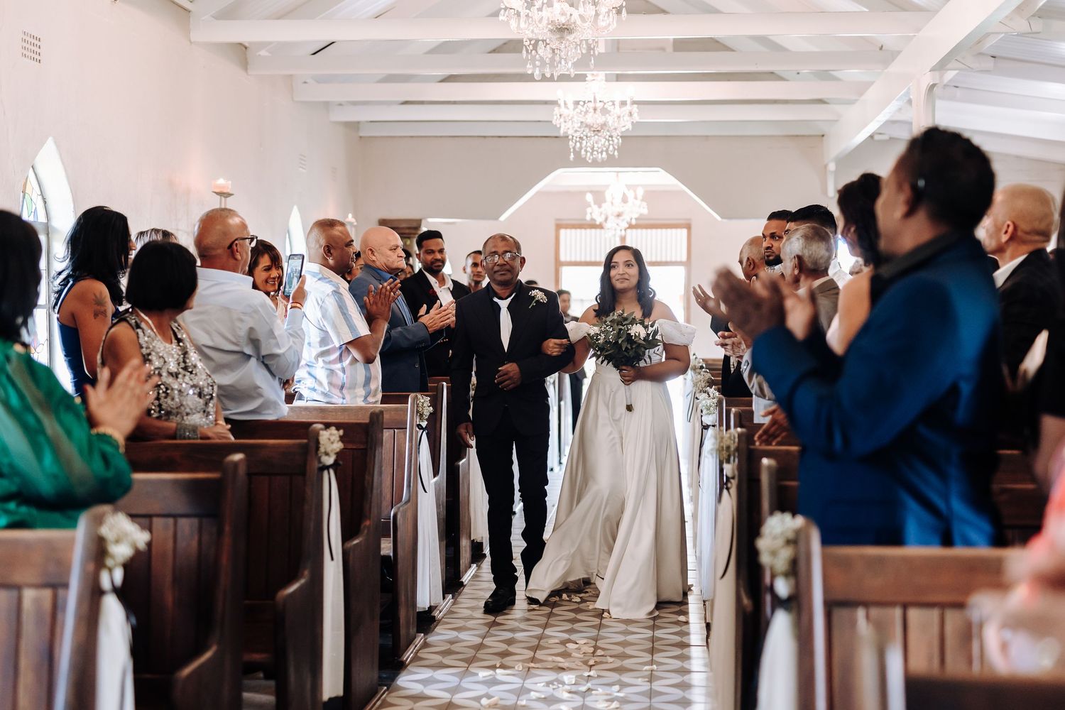 Wedding guests applaud as the newlyweds walk down the aisle in a chapel with chandeliers.