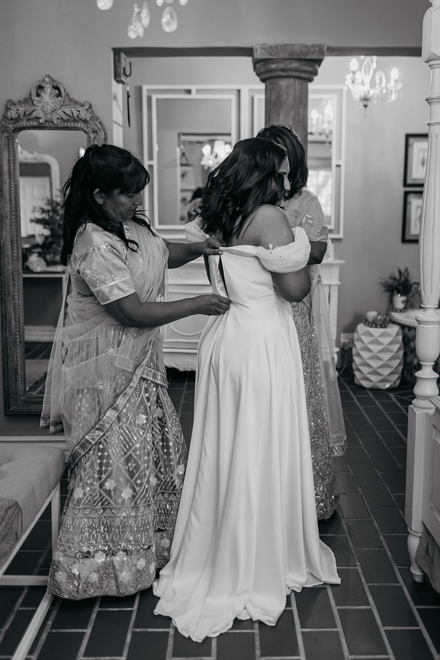 Wedding preparation scene showing dress fitting in elegant room with chandelier and ornate mirror.