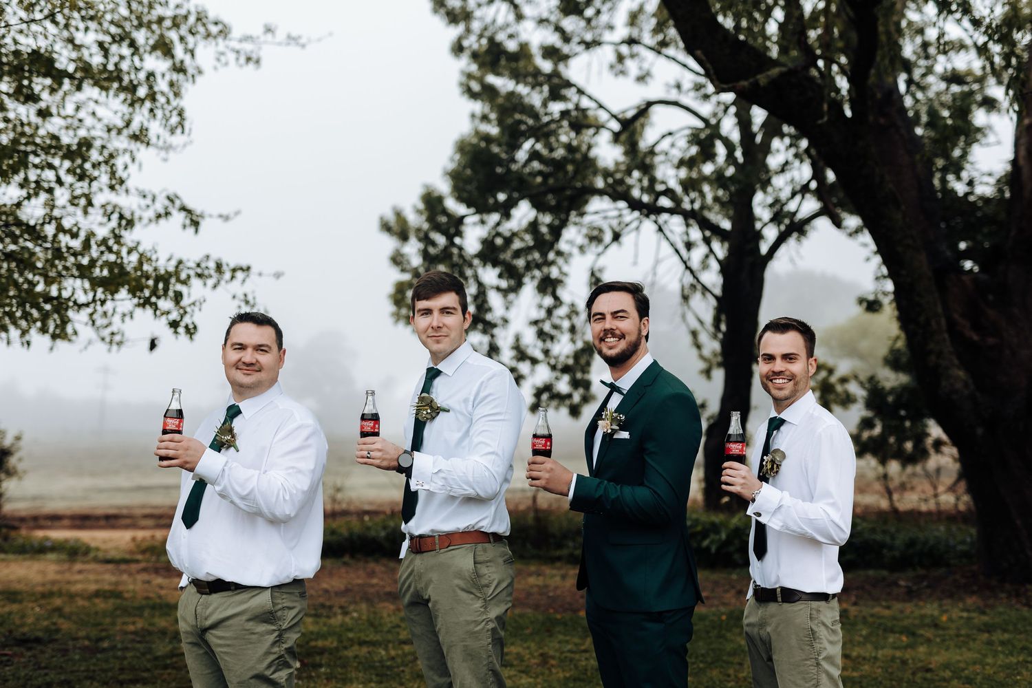 Wedding party holds up beer bottles in a casual outdoor photo against a misty landscape.