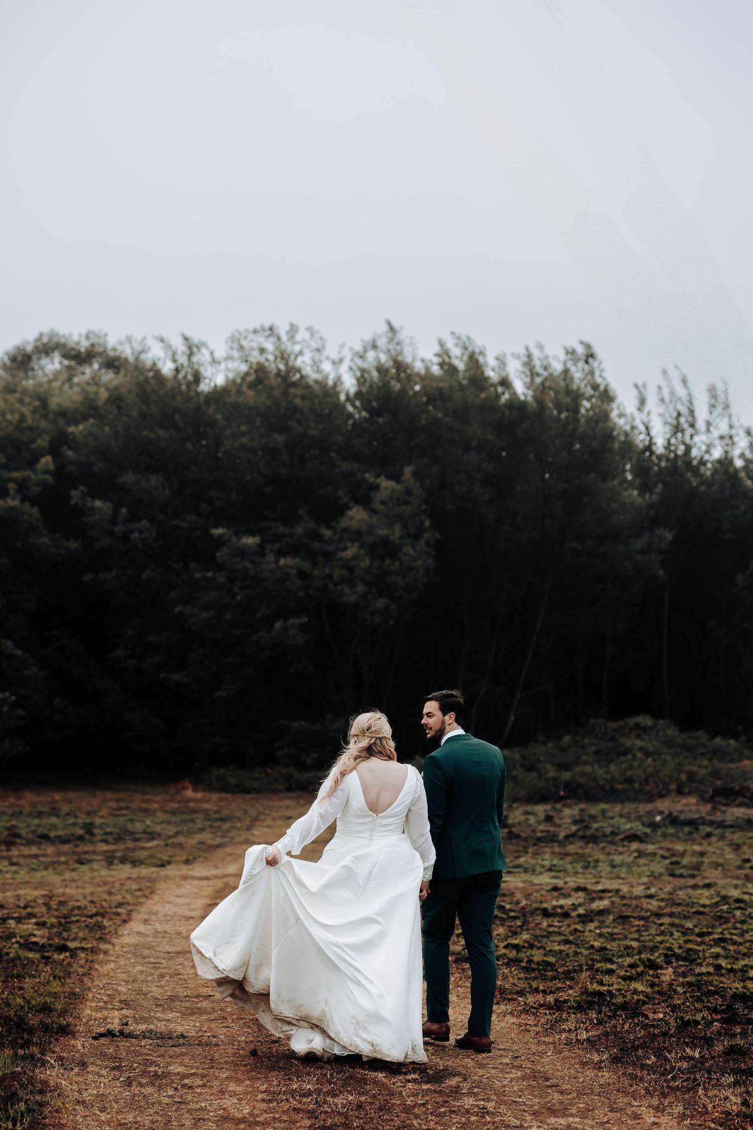 Newlyweds walk together on a forest trail as the bride's dress flows behind them.