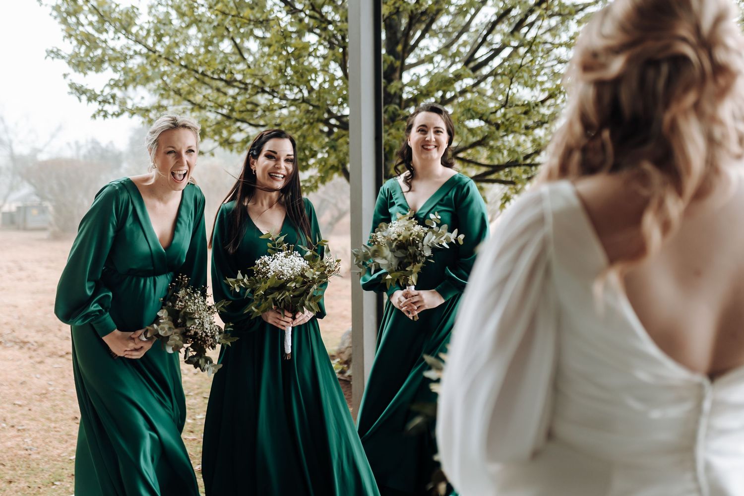 Bridesmaids in emerald green dresses holding bouquets outdoors with bride in foreground.