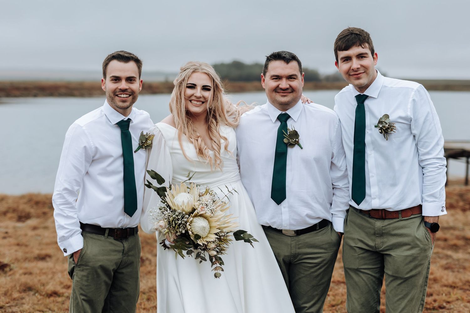 Wedding party in white shirts and green ties poses together against overcast lakeside backdrop.