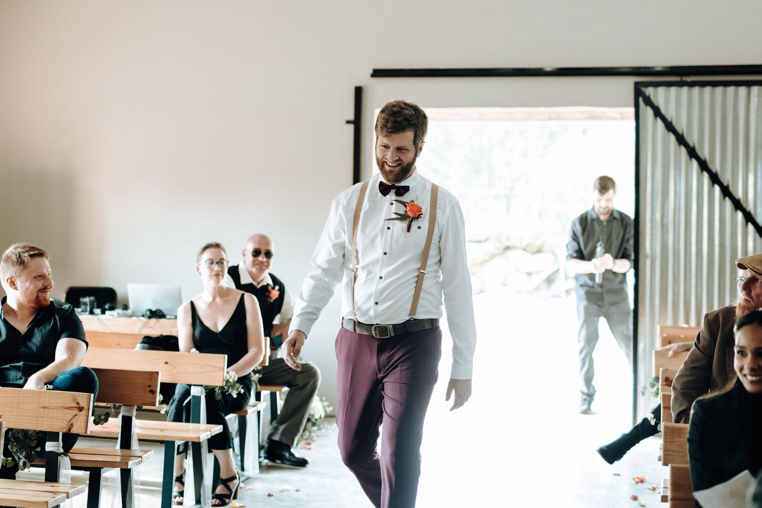 Wedding party member in burgundy pants and suspenders walks through bright indoor venue.