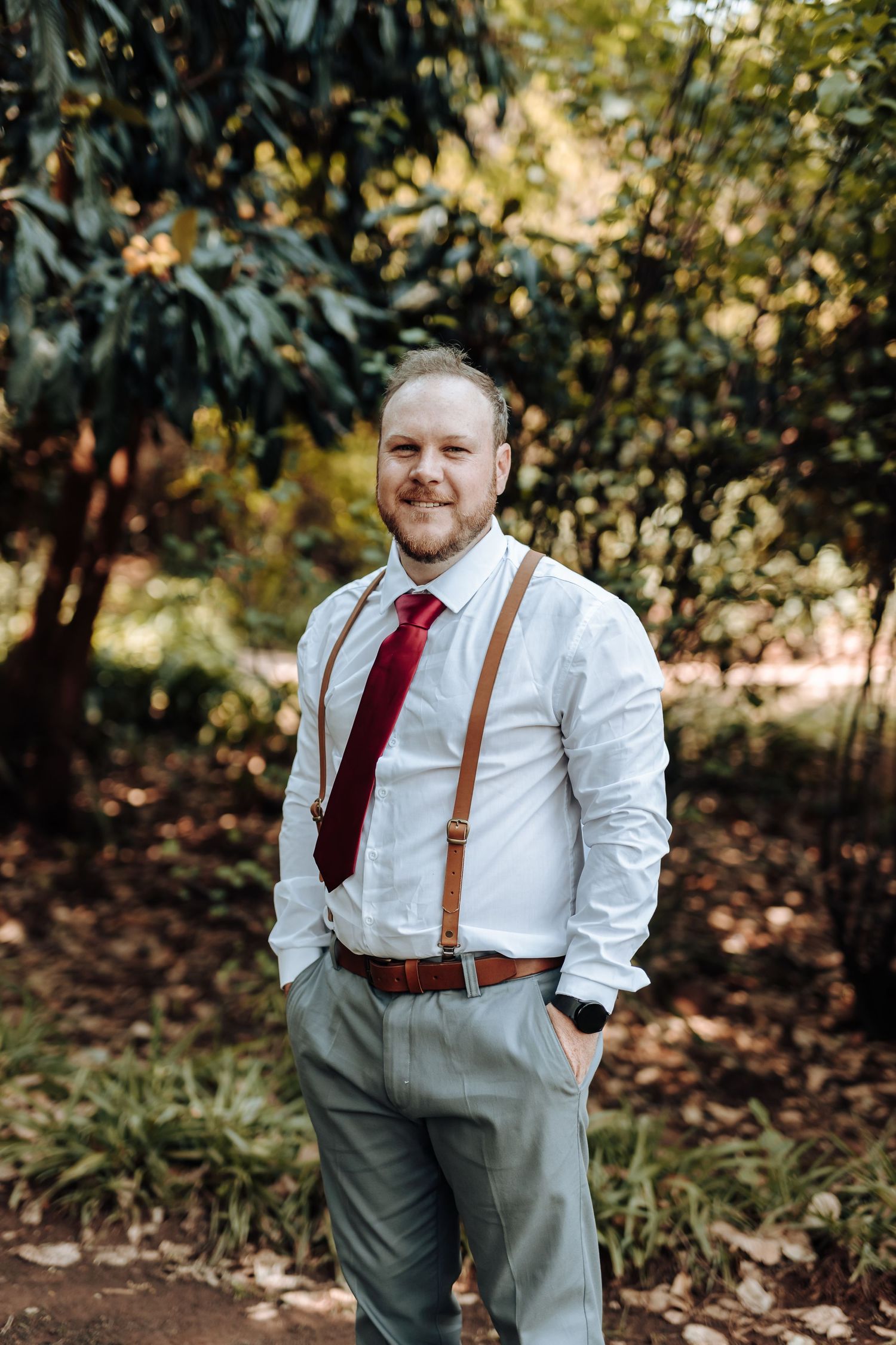 Man in formal attire with suspenders poses casually in outdoor setting.