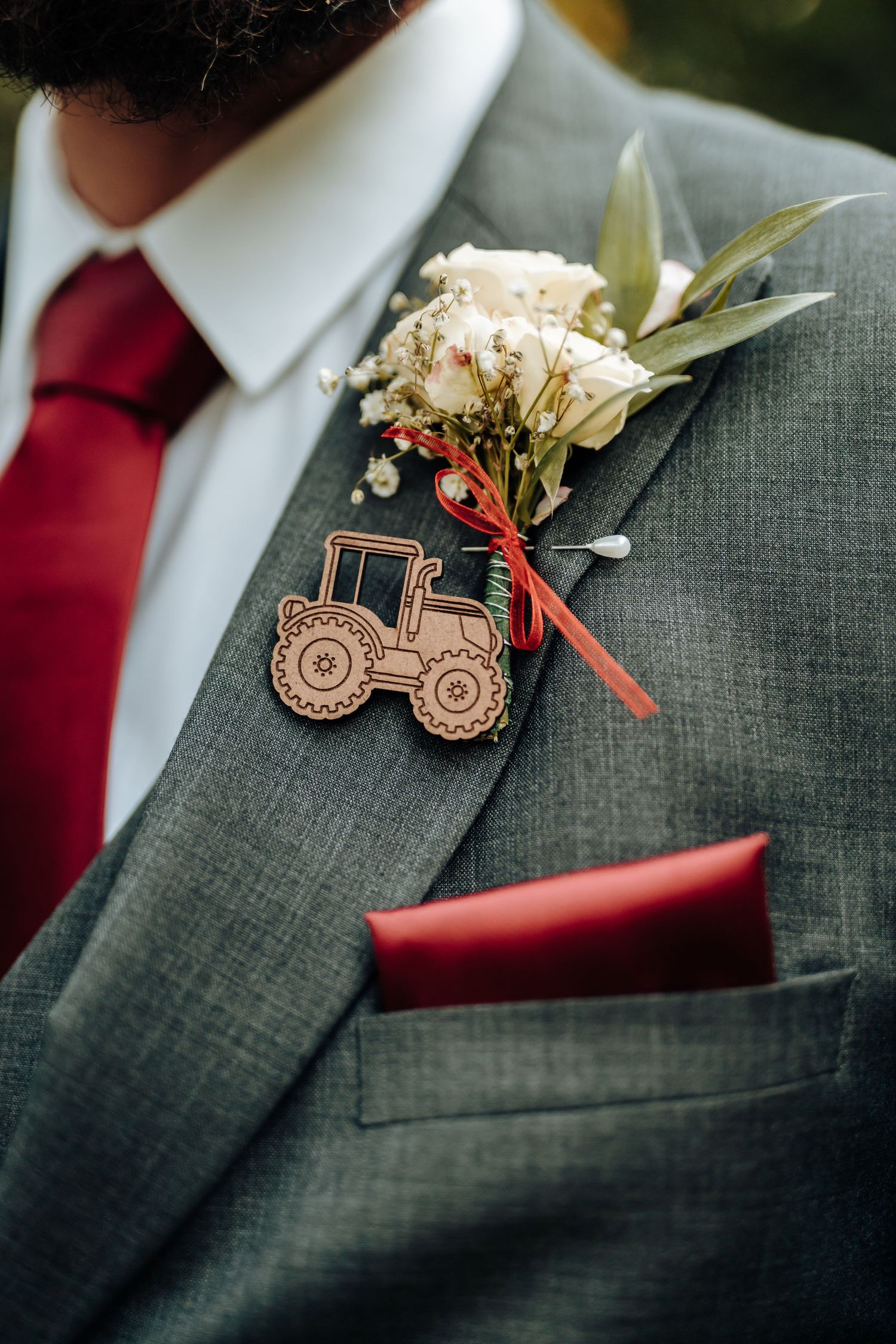 Close-up of wooden tractor boutonniere and white flowers on grey suit lapel with red tie.