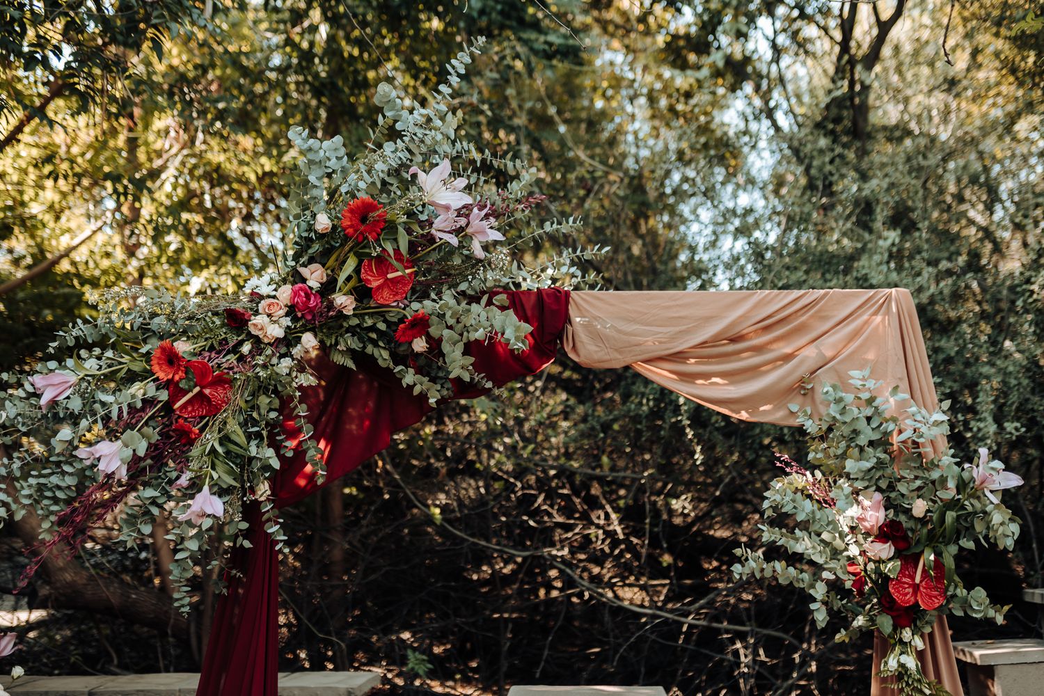 Outdoor wedding arch decorated with burgundy flowers and blush fabric in a garden setting.