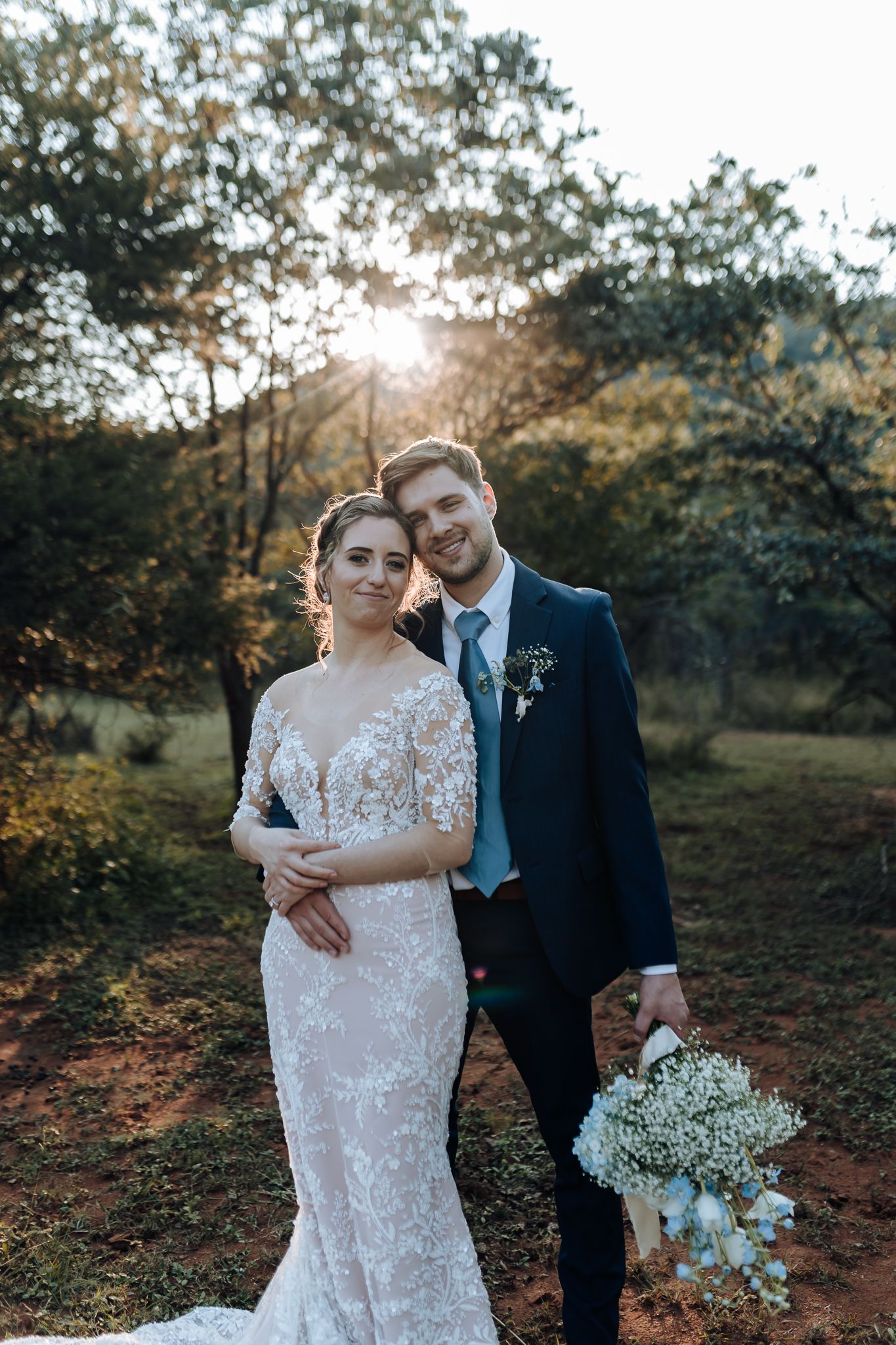 Sunset wedding portrait with couple standing together among trees while holding delicate white bouquet.