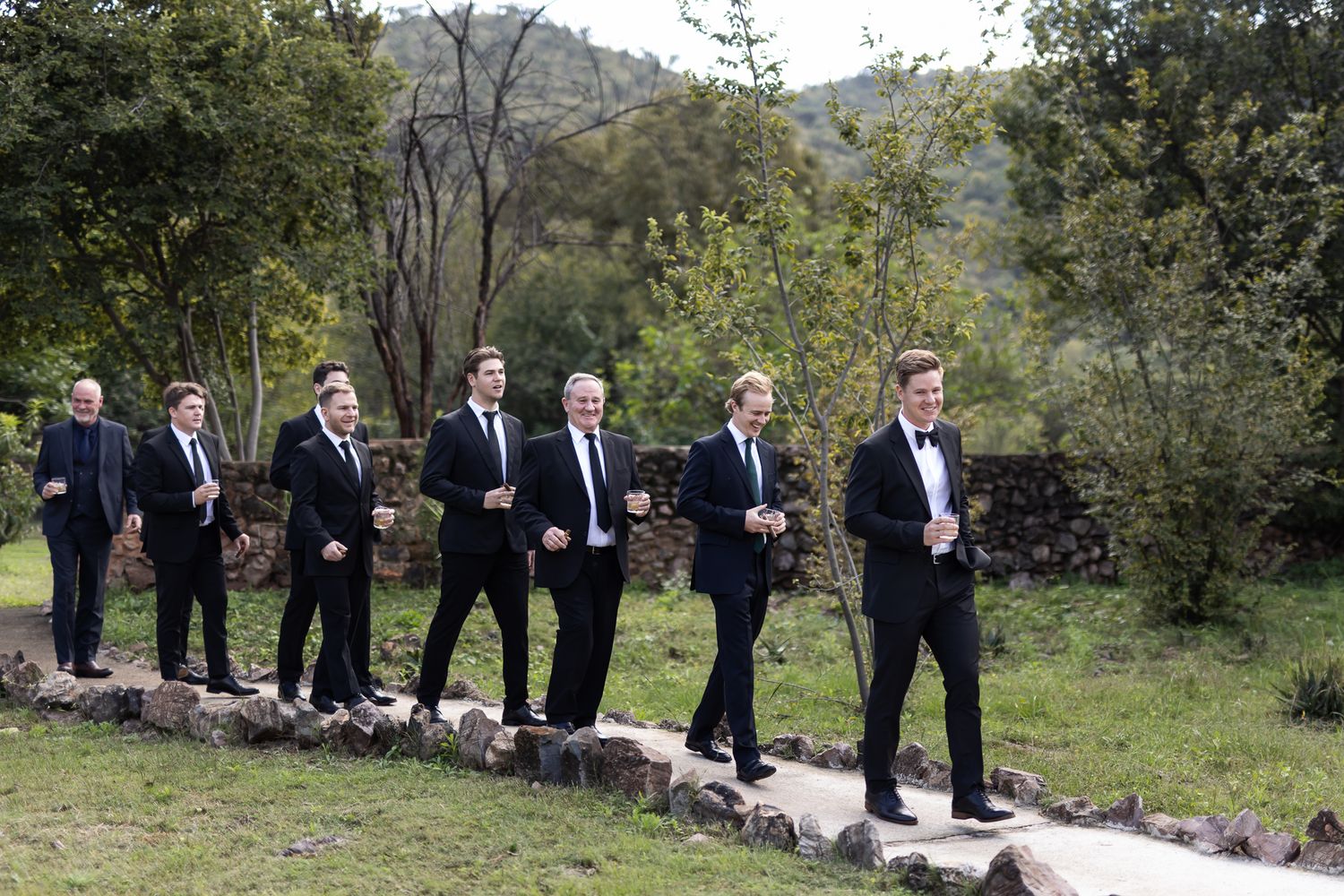 Wedding party in dark suits walking along stone path in natural setting.