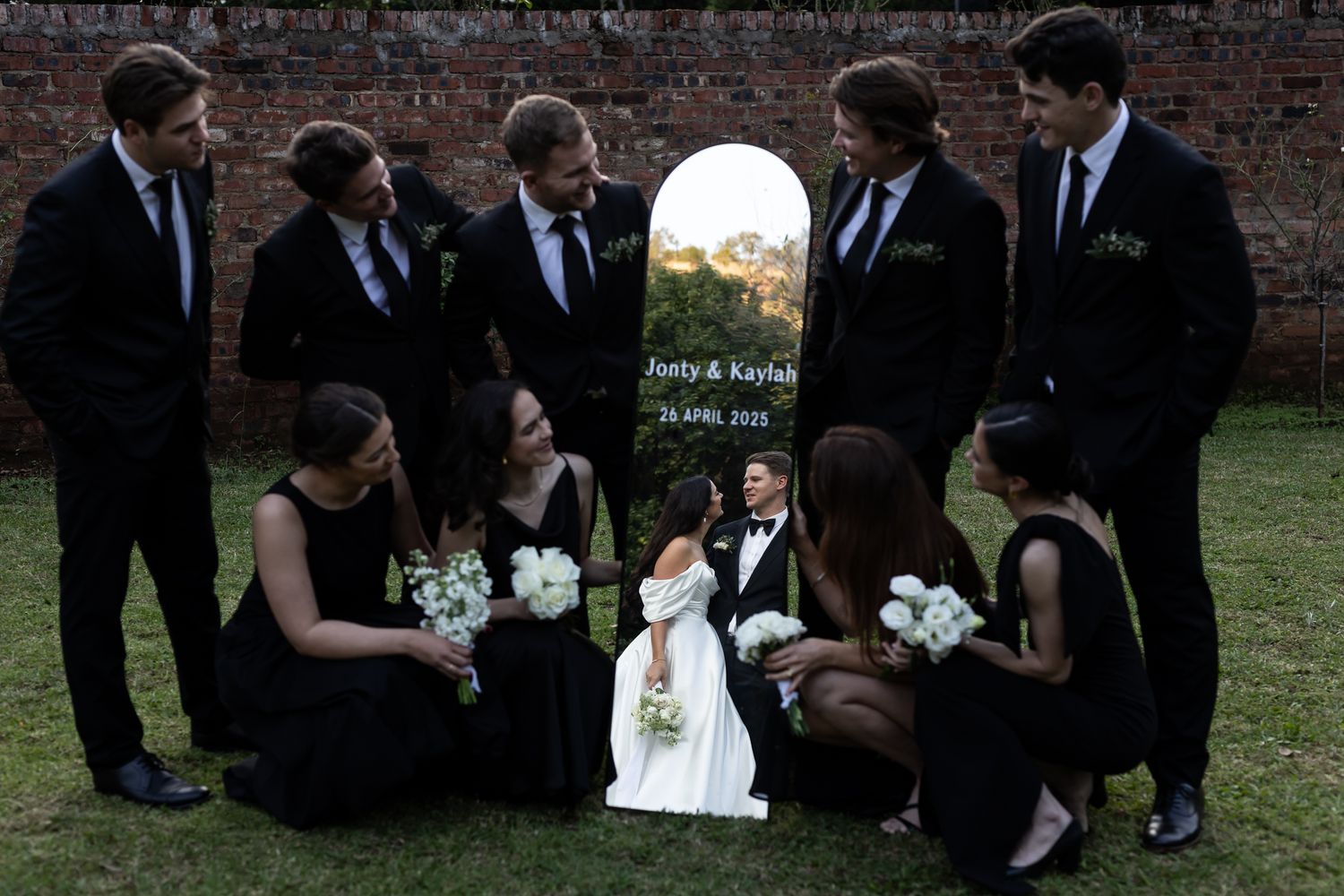 Wedding party in black attire gathered around small child in white dress against brick wall backdrop.