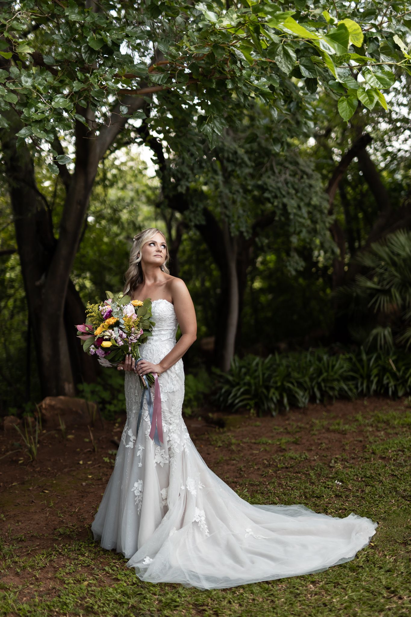 A full-length bridal portrait in a lush garden setting with a flowing lace gown and trailing bouquet.