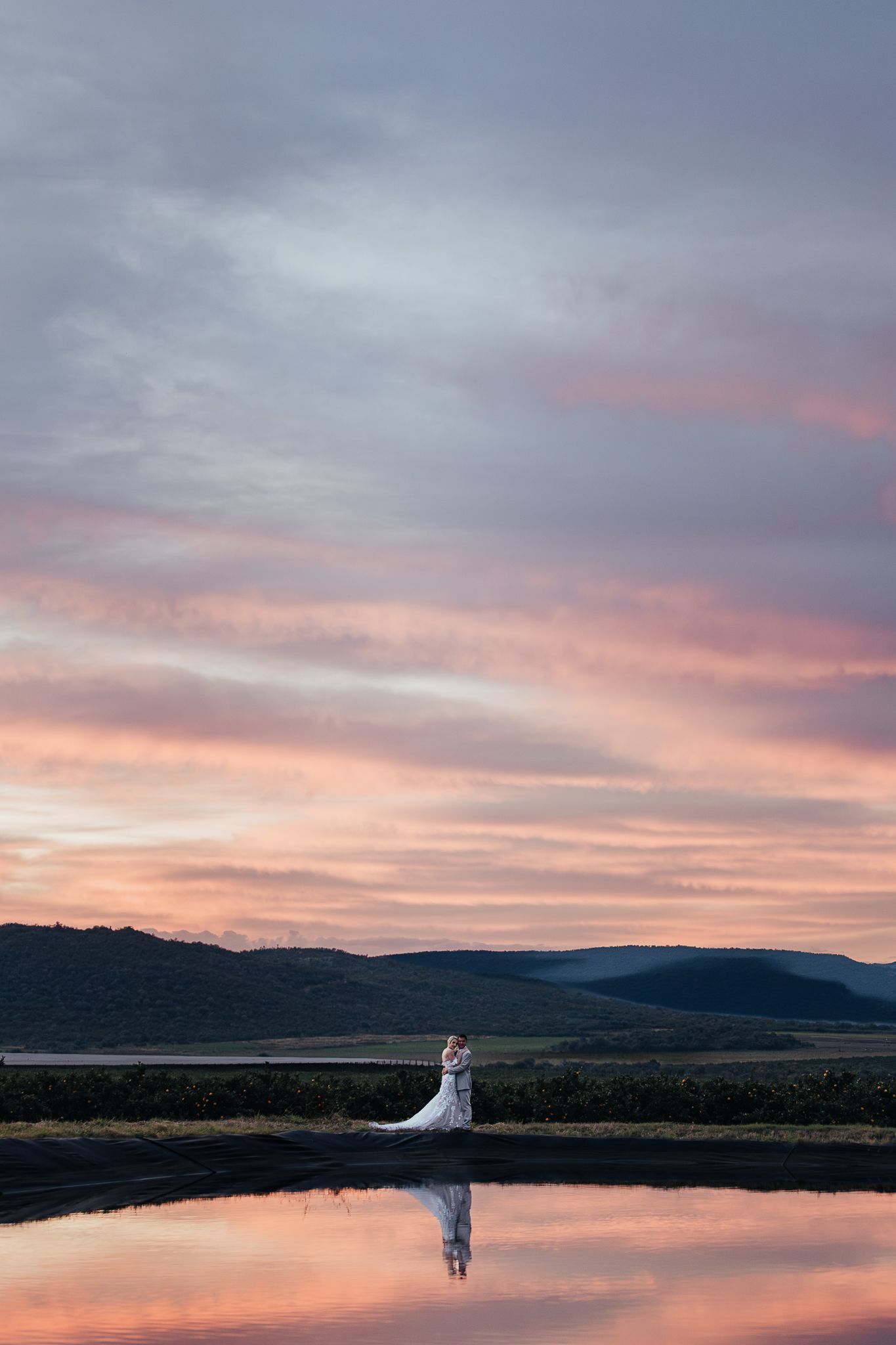Dramatic sunset wedding photo reflected in still water with mountains silhouetted in background.
