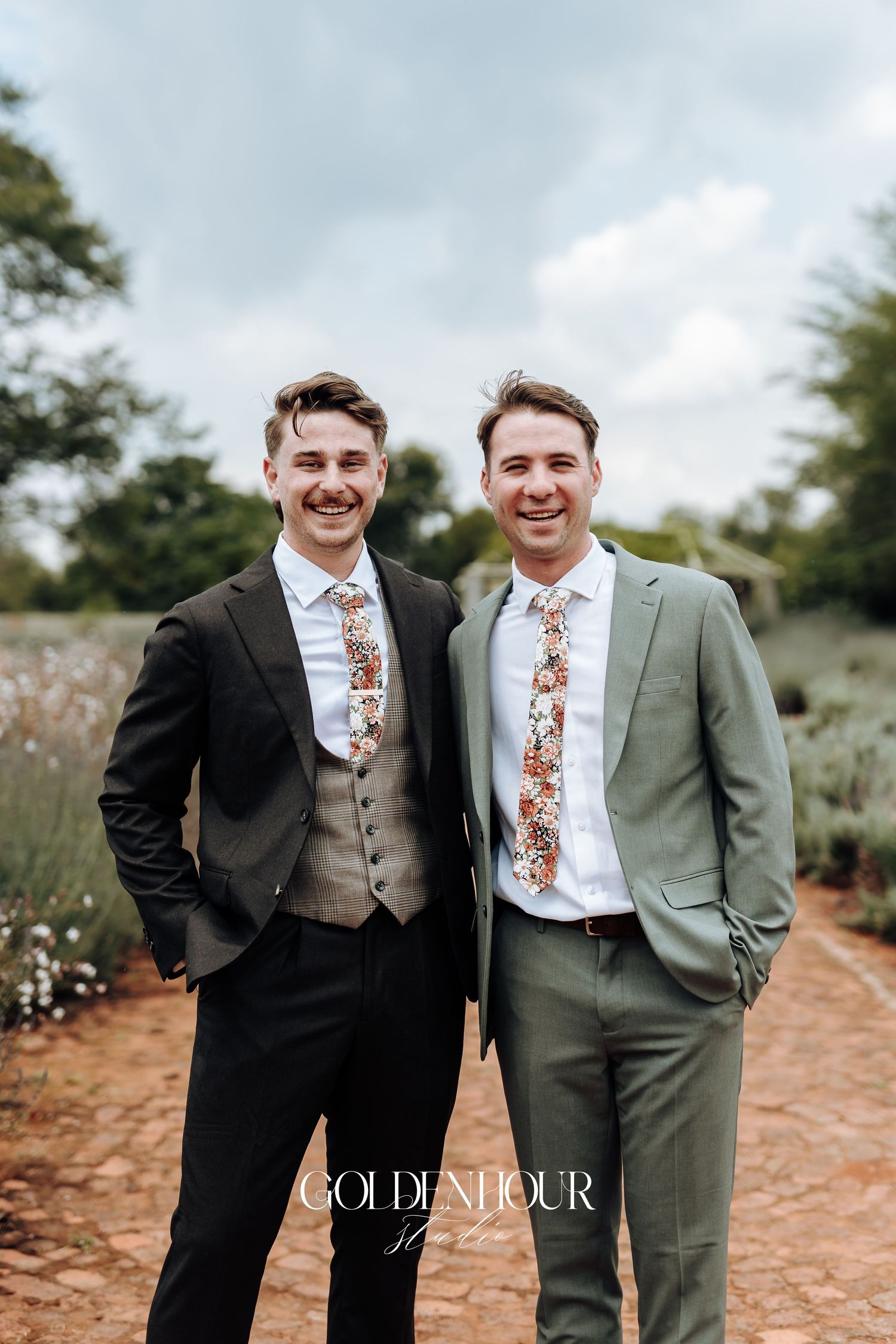 Two people in suits with floral ties pose together outdoors against a natural backdrop.