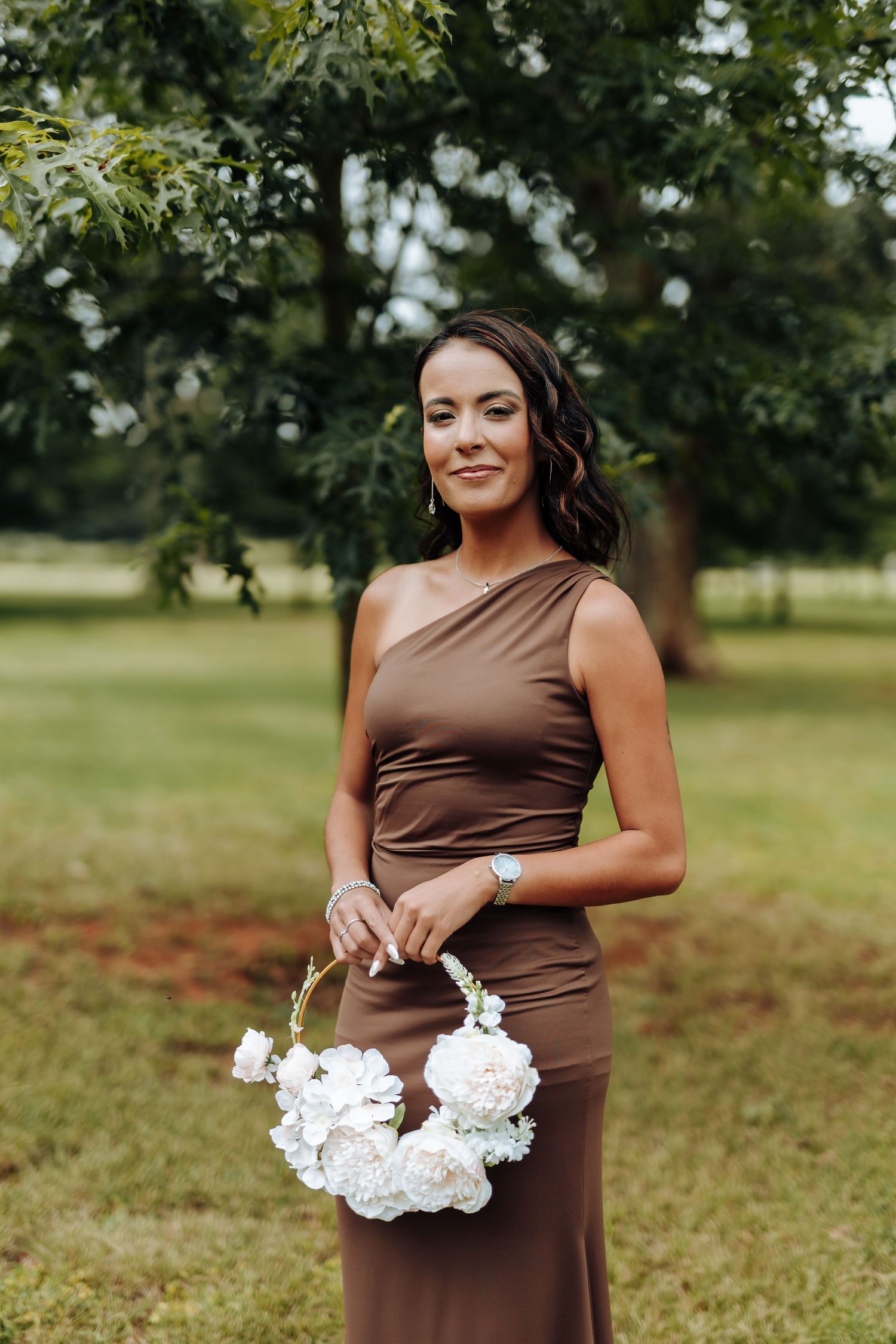 A bridesmaid in a brown dress holds a white bouquet while posing in a grassy outdoor setting.