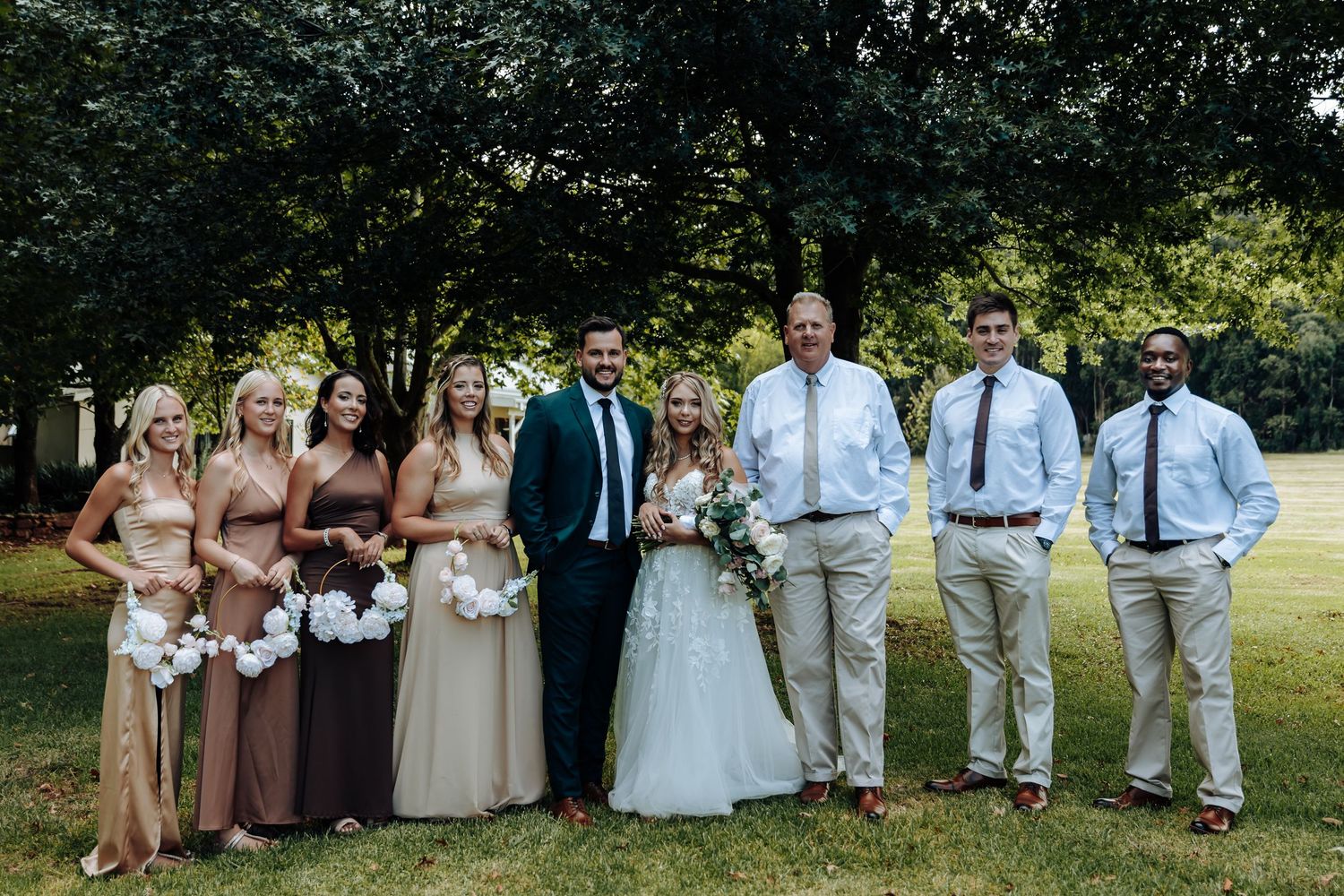 A wedding party poses together outdoors in a garden setting wearing formal attire and holding bouquets.