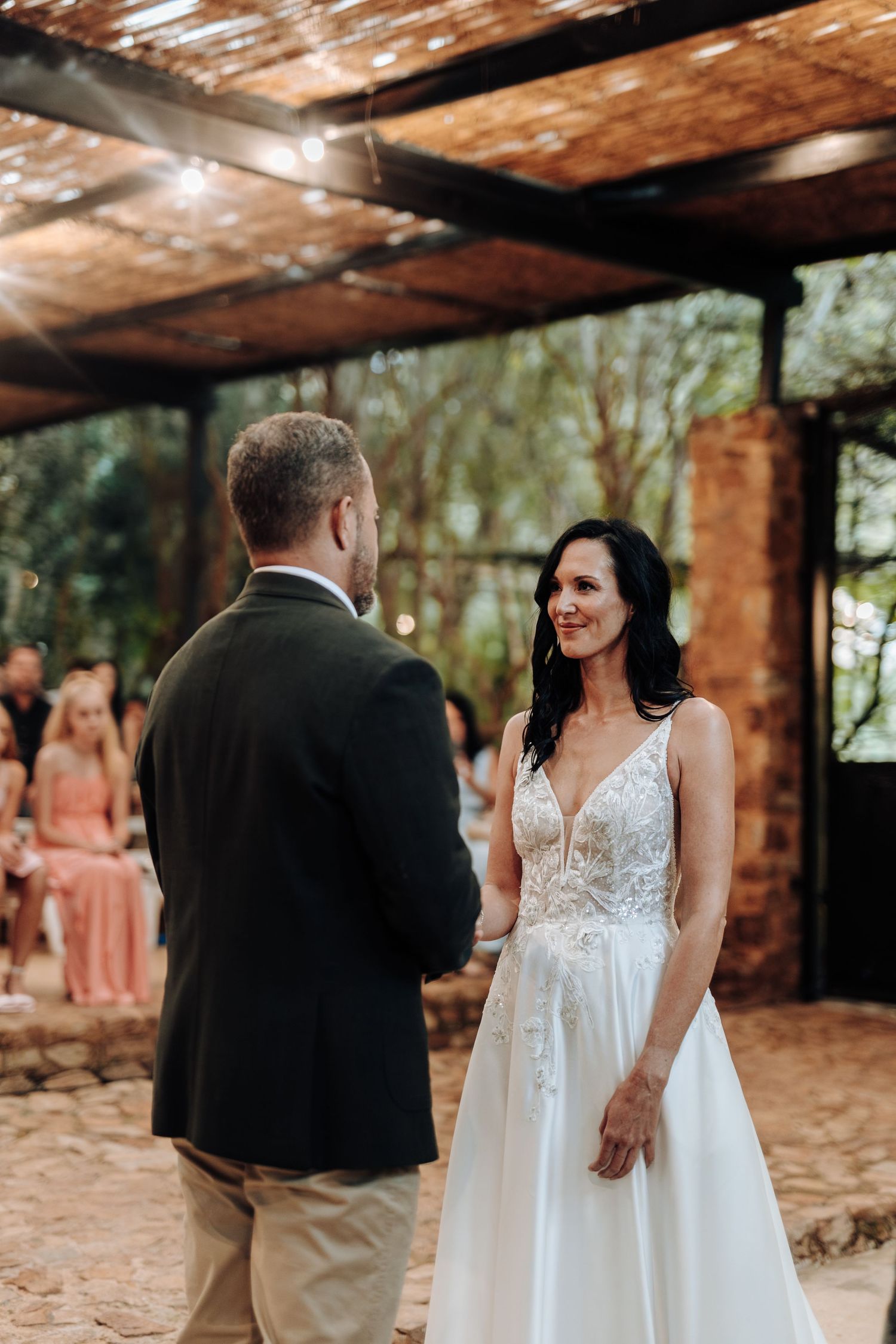 Bride in white lace gown smiling at groom during outdoor wedding ceremony under rustic wooden pergola.