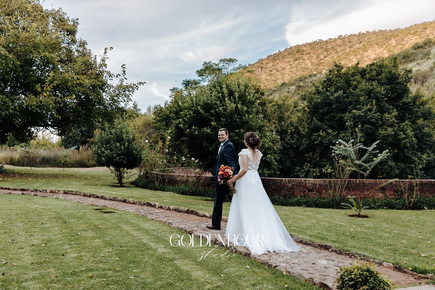 Bride and groom standing in lush garden with mountain backdrop, bride holding vibrant red bouquet.