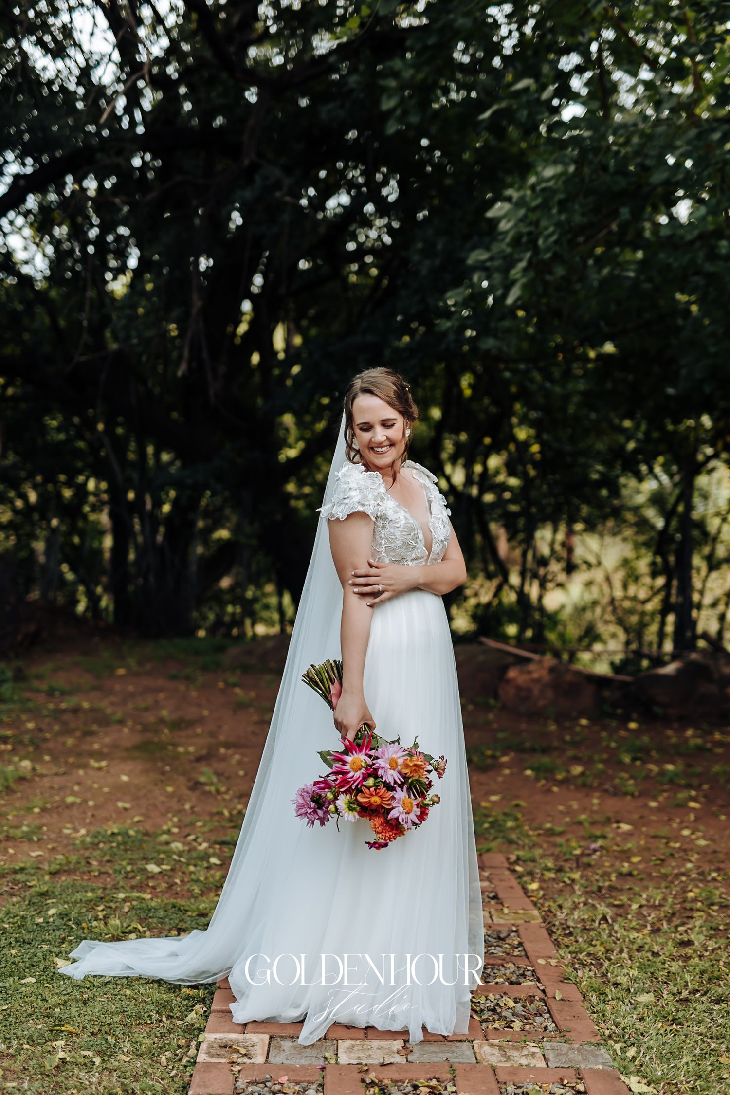 Elegant bride in lace wedding gown holding colorful bouquet on brick path among trees.