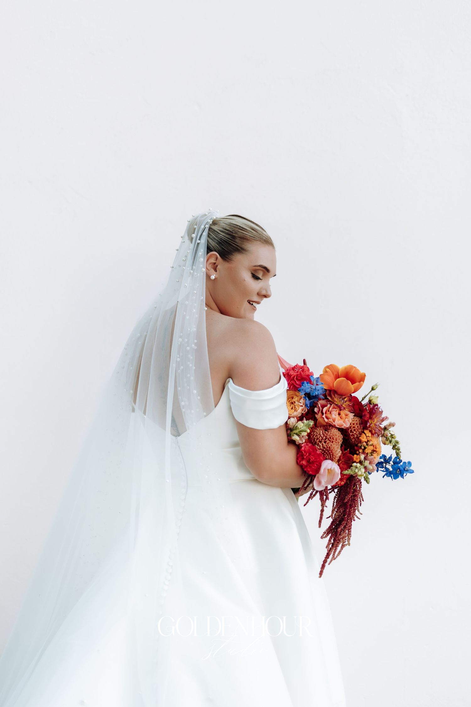 Side view of bride in white gown with colorful bouquet featuring red, orange and blue flowers against white background.