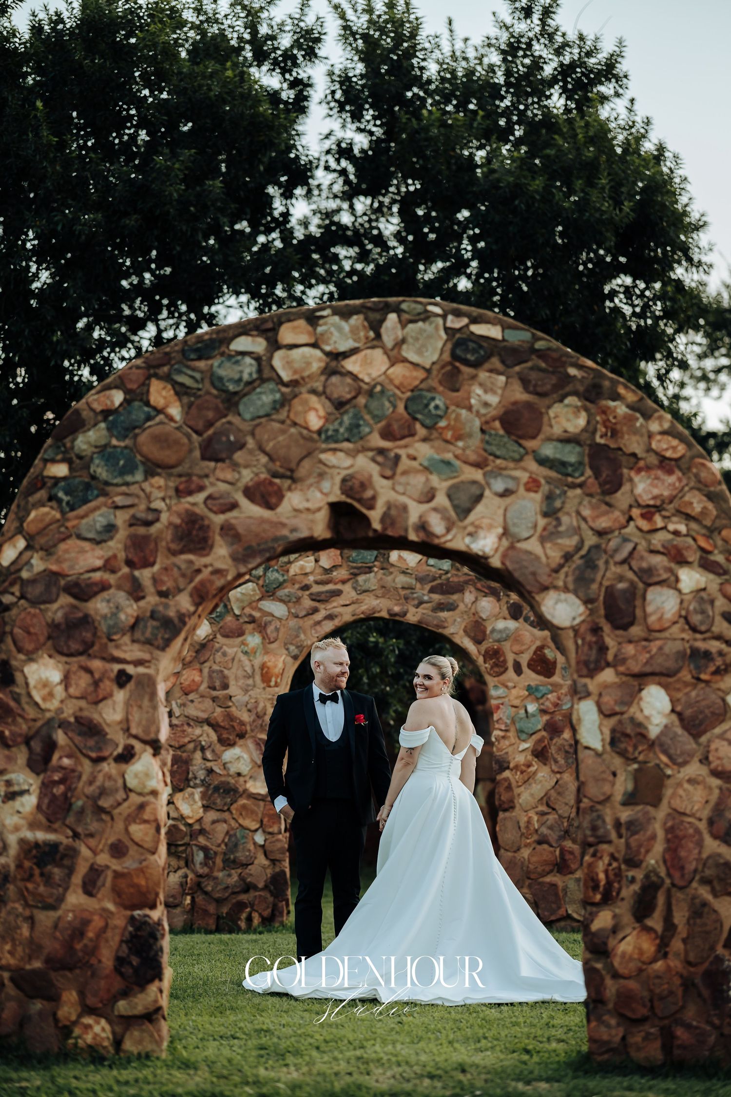 A stone archway in a garden setting provides a romantic backdrop for a formal wedding portrait.