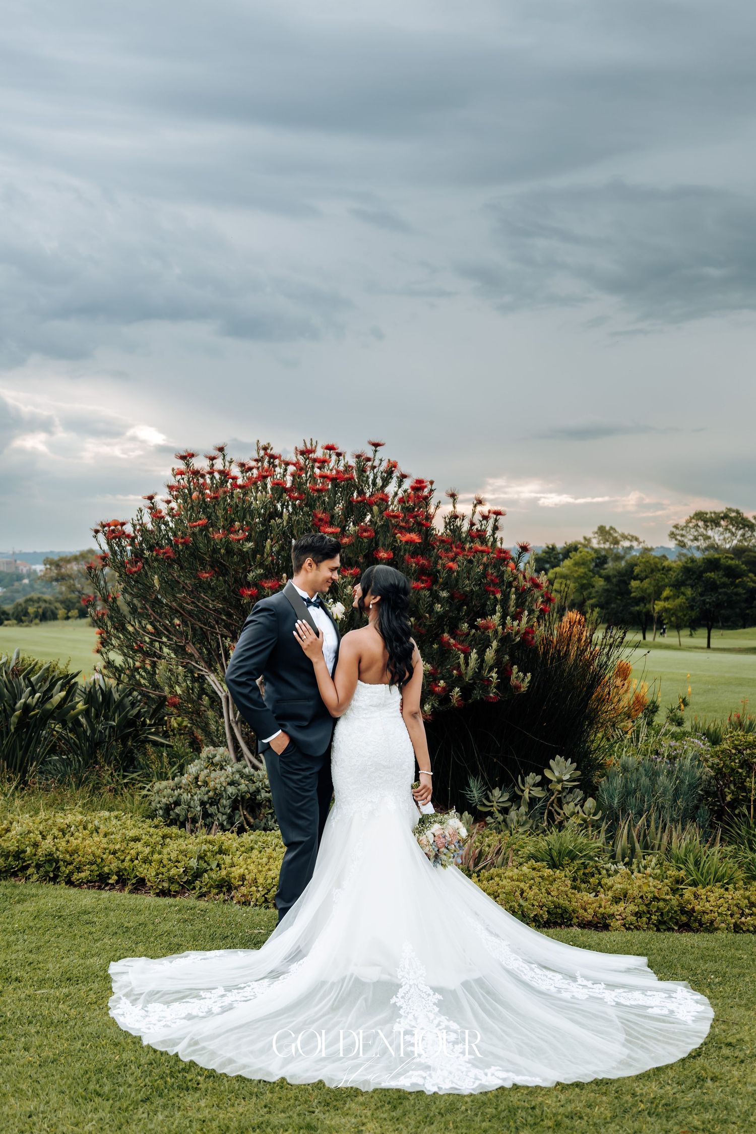 Wedding couple poses in front of red flowering bush with dramatic train on white dress.
