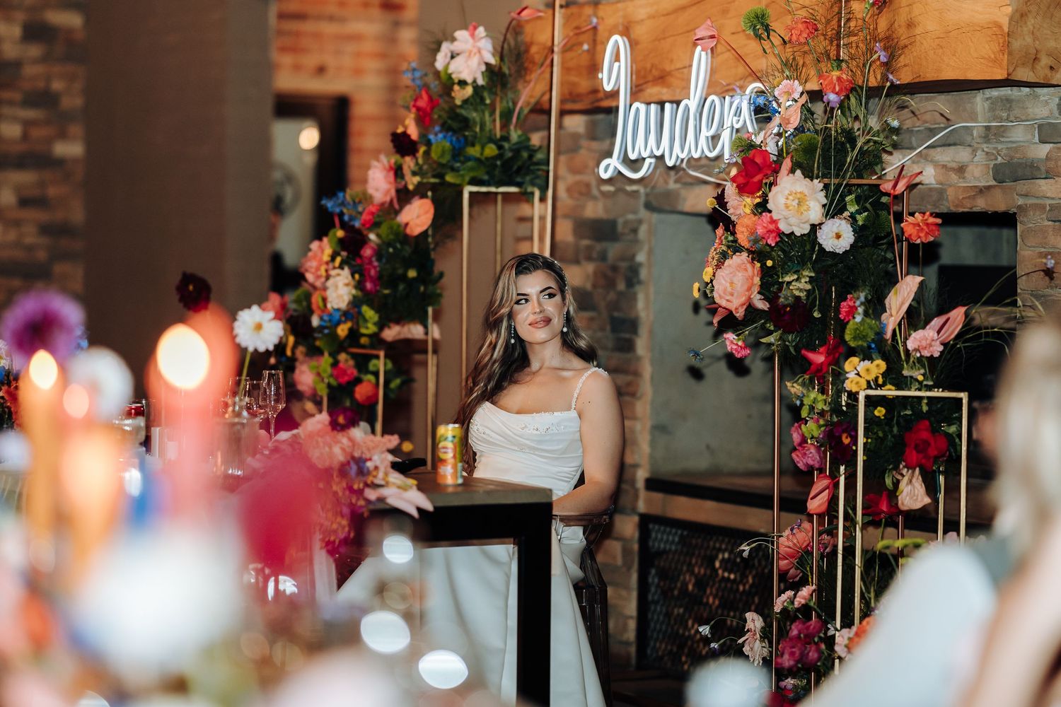 Wedding reception setup with floral arrangements and neon sign against rustic brick wall.