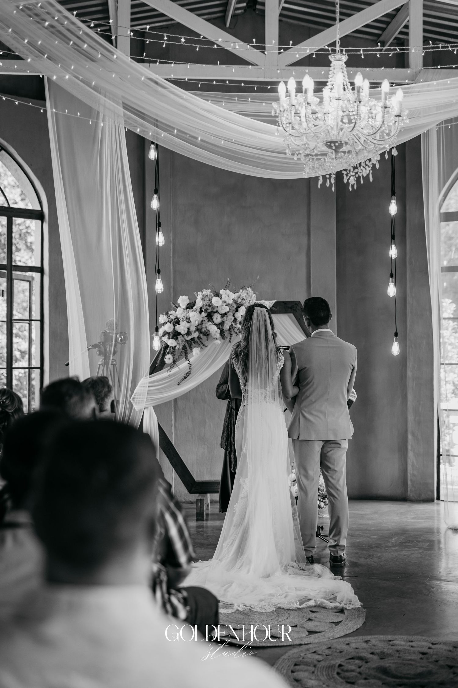 Black and white photo of a wedding ceremony in an elegant ballroom with crystal chandeliers and draped fabric.