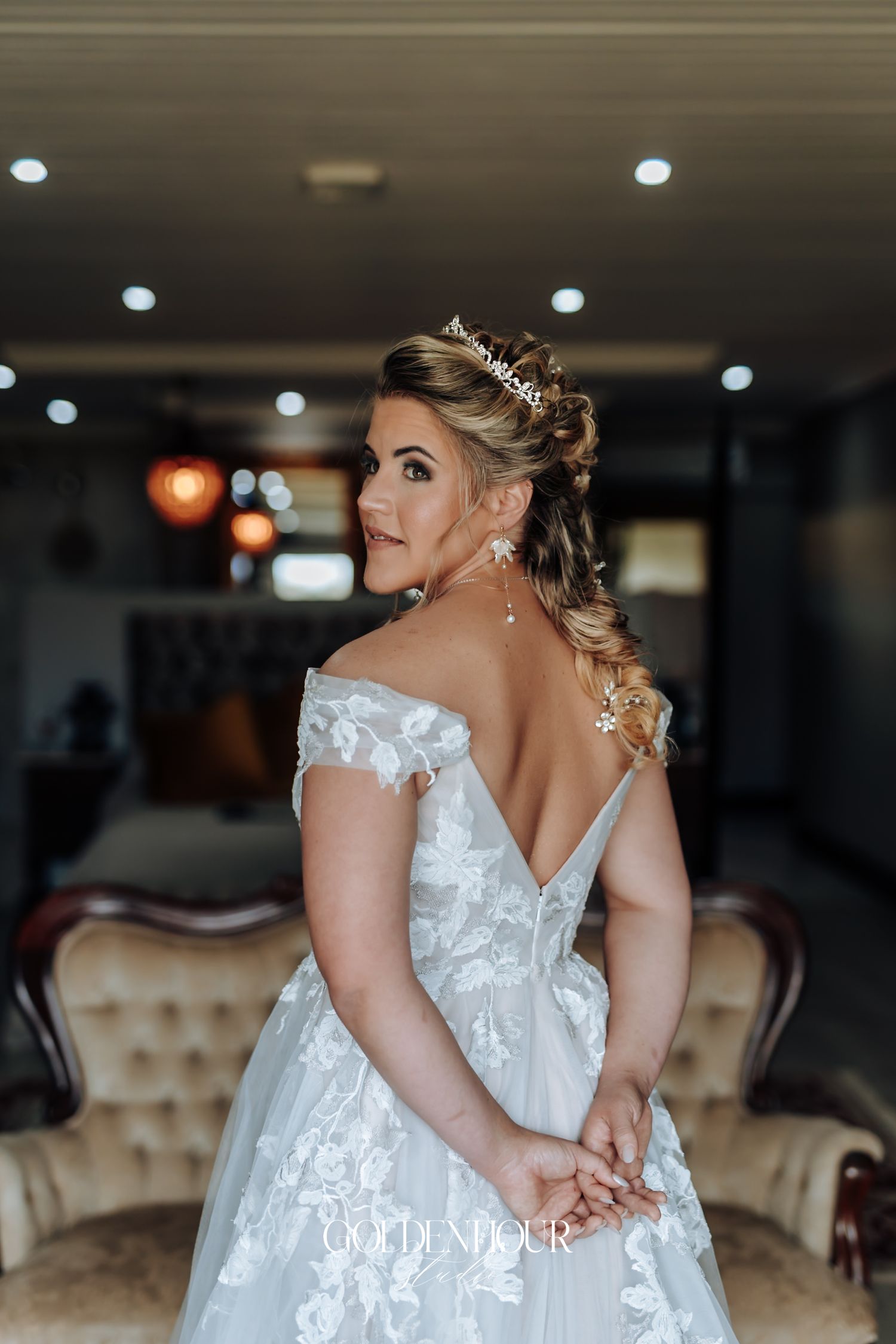 Elegant bride in off-shoulder lace wedding gown and tiara poses looking over shoulder indoors.