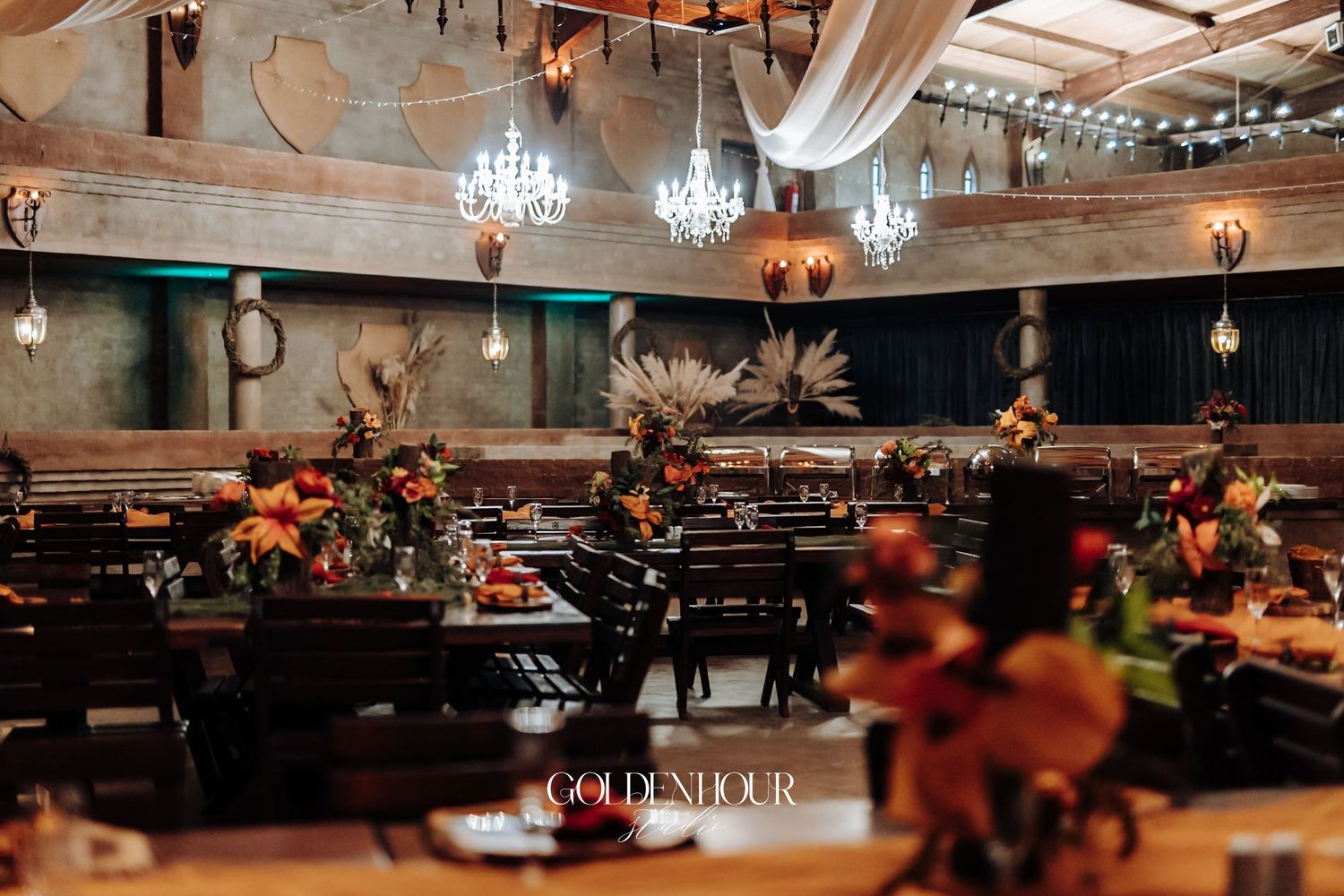 Elegant wedding reception venue with chandelier lighting, dark wood tables, and vibrant floral centerpieces in warm autumn tones.