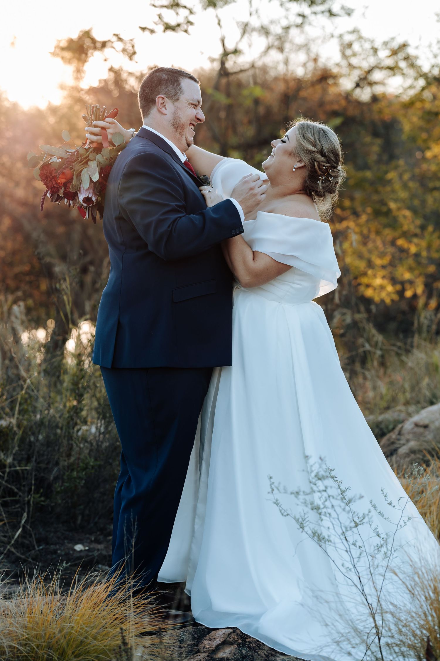 Bride and groom share a joyful embrace outdoors at golden hour, she in white off-shoulder gown, he in navy suit.