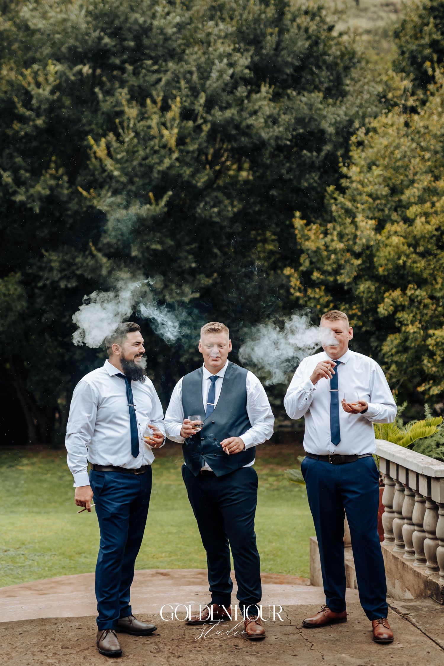 Three groomsmen in navy pants and white shirts smoking cigars outdoors at a wedding celebration.