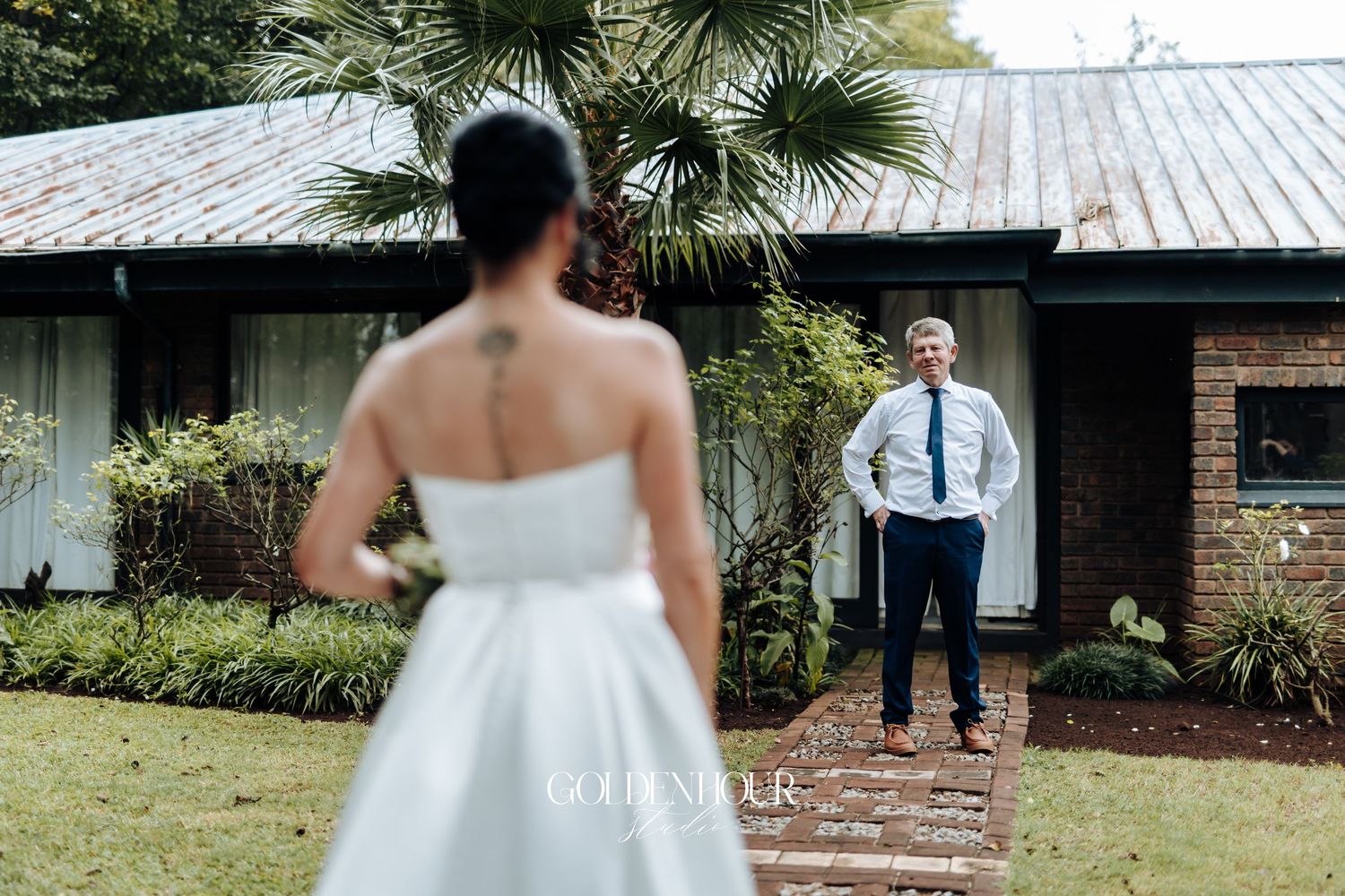 Groom stands in garden watching bride approach in white strapless gown during outdoor wedding first look.