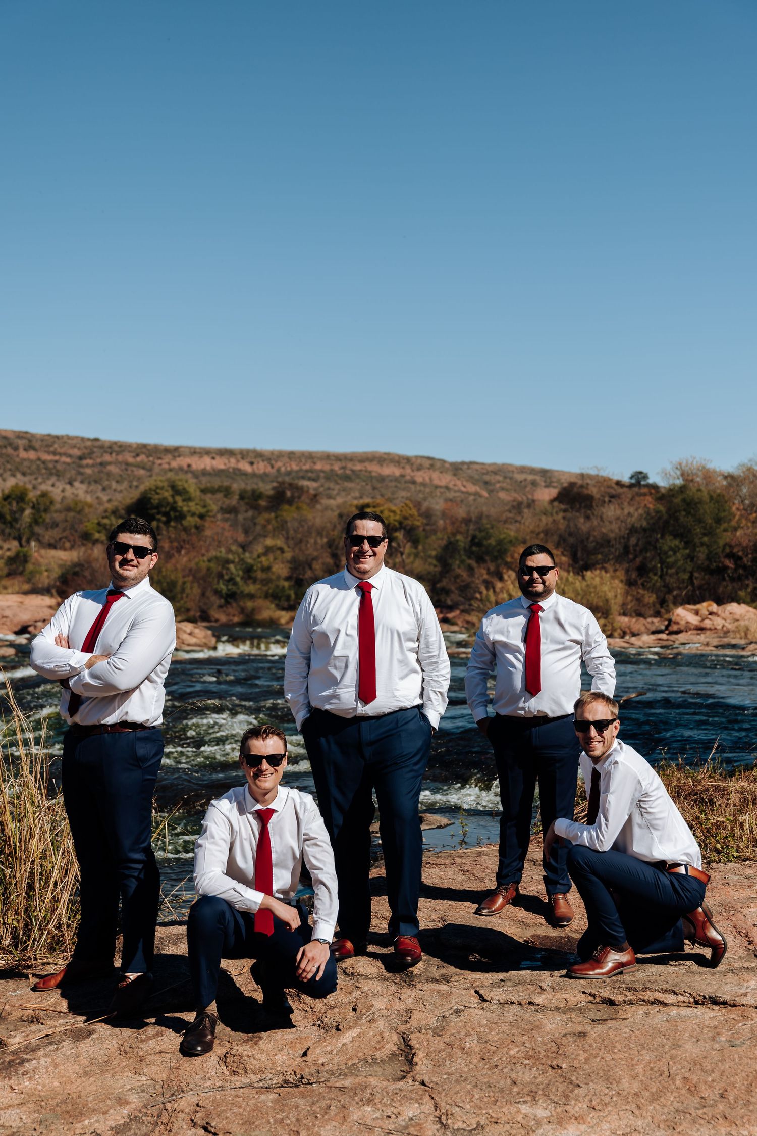 Five groomsmen in white shirts and red ties pose on rocks by a river under a clear blue sky.