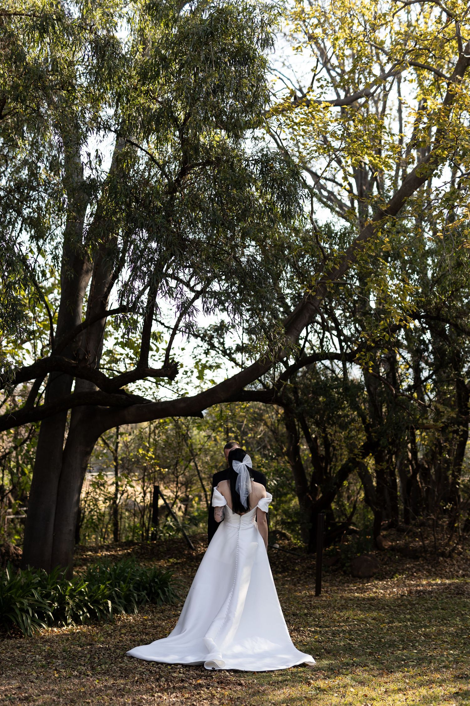 A figure in a flowing white wedding dress stands facing tall eucalyptus trees in a serene outdoor garden setting.