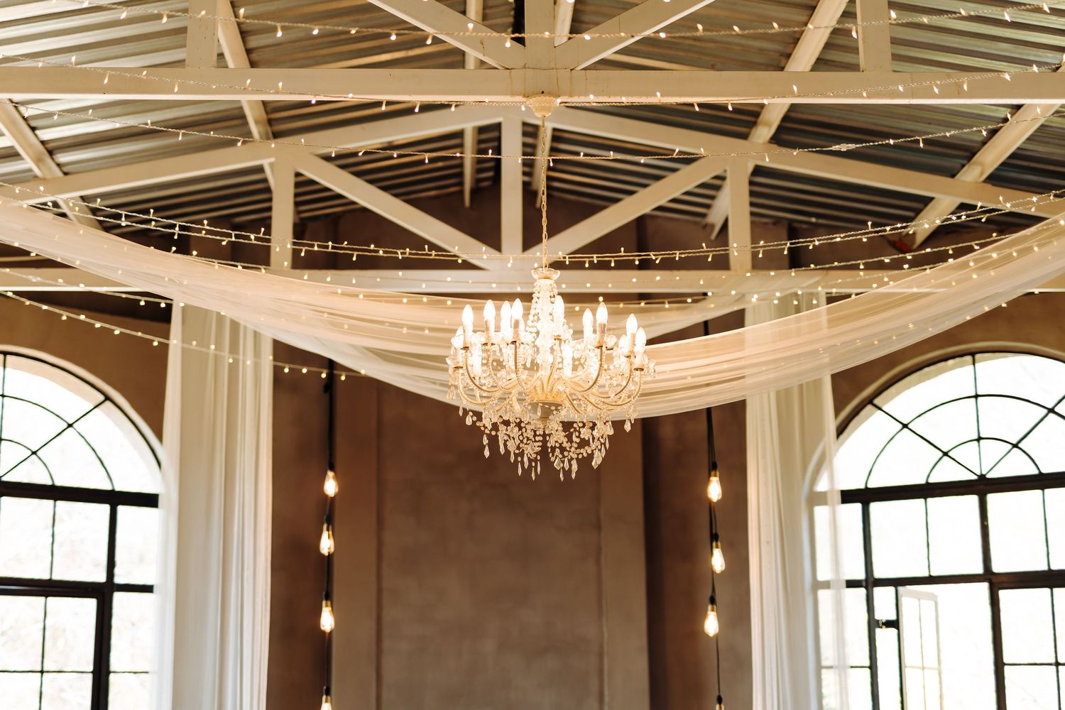 Wedding reception venue with exposed wooden beams, chandeliers and white fabric draping across elegant ballroom ceiling.
