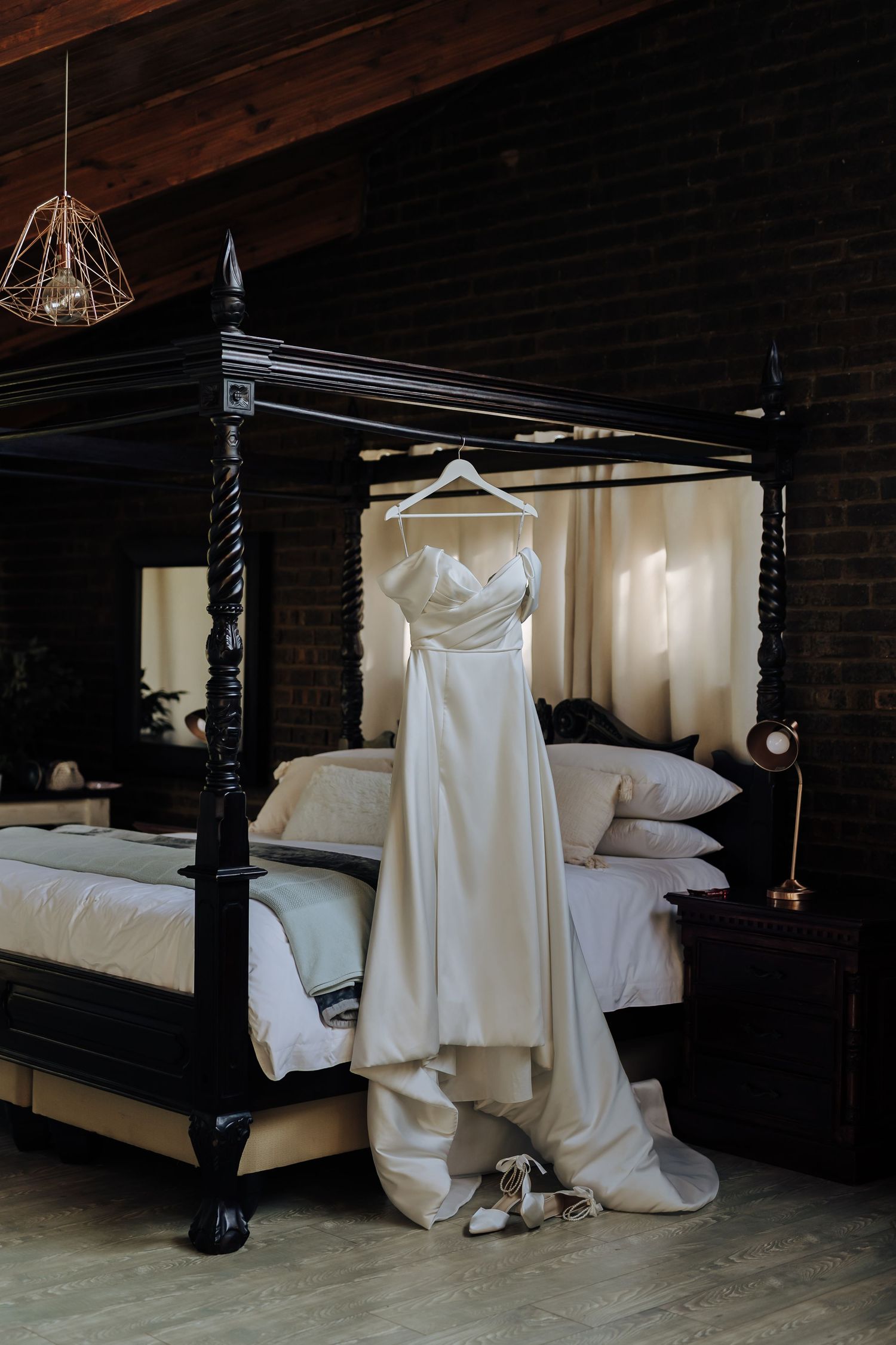 An elegant white wedding dress hangs on a black four-poster bed with silver heels beneath in a moody bedroom setting.
