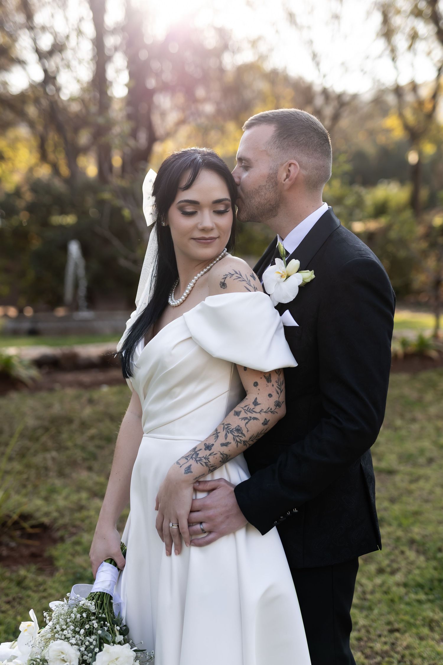 Couple in wedding attire embraces outdoors during ceremony, with bride in white dress showing intricate arm tattoos.