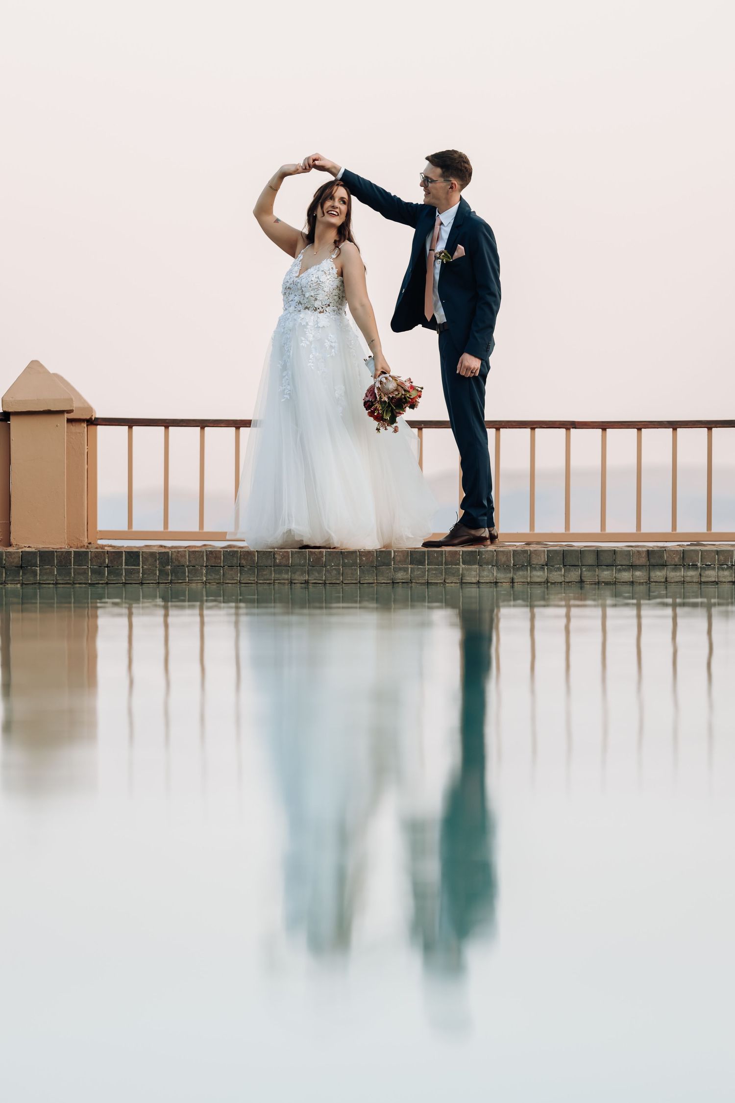 A couple in wedding attire dance together on a balcony with a reflection in the foreground.