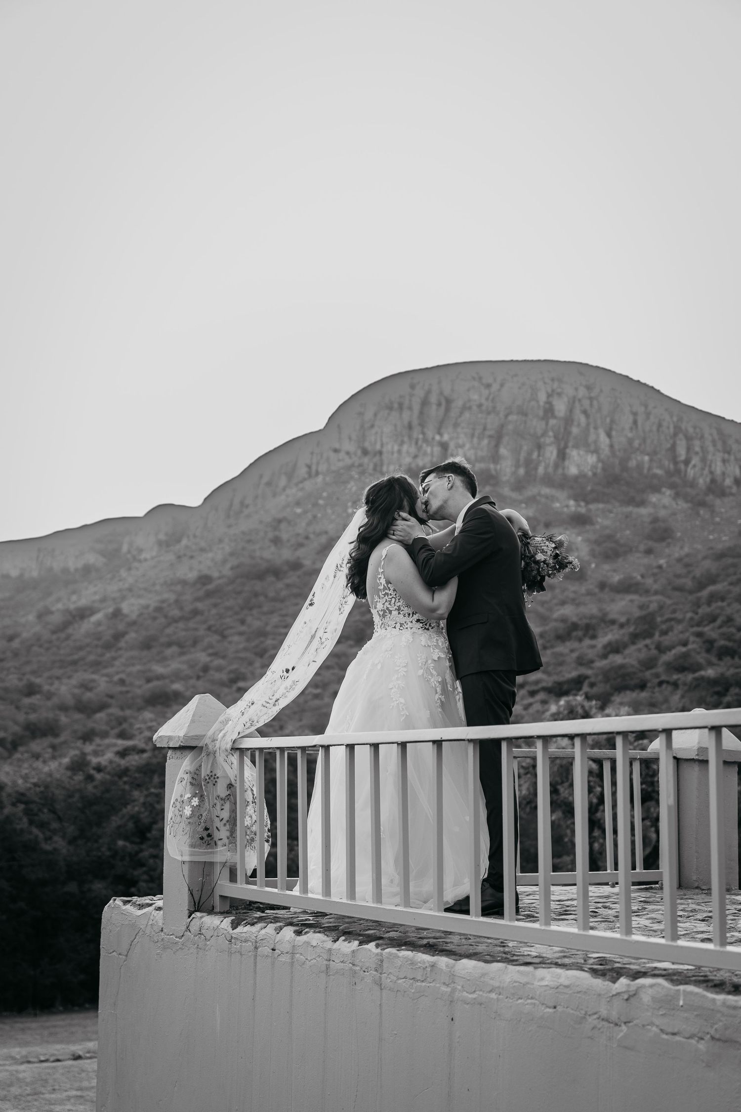 A romantic black and white wedding photo showing a couple sharing a kiss on a balcony with mountains in the background.
