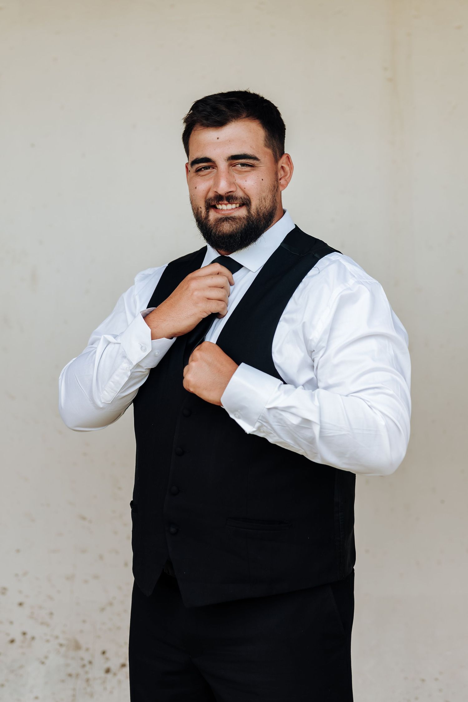 A person in a white dress shirt and black vest laughs while adjusting their collar against a light background.