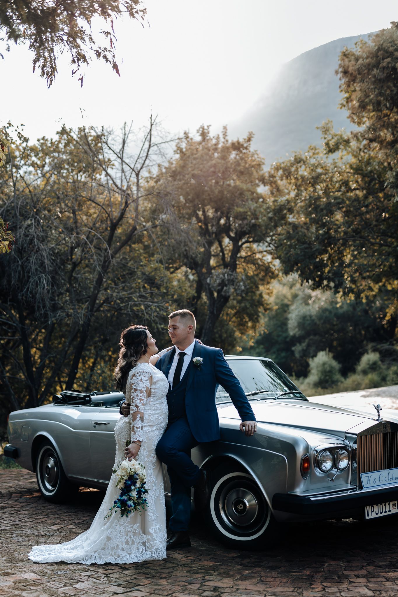 Elegant bride and groom pose with vintage Rolls Royce against mountain landscape.